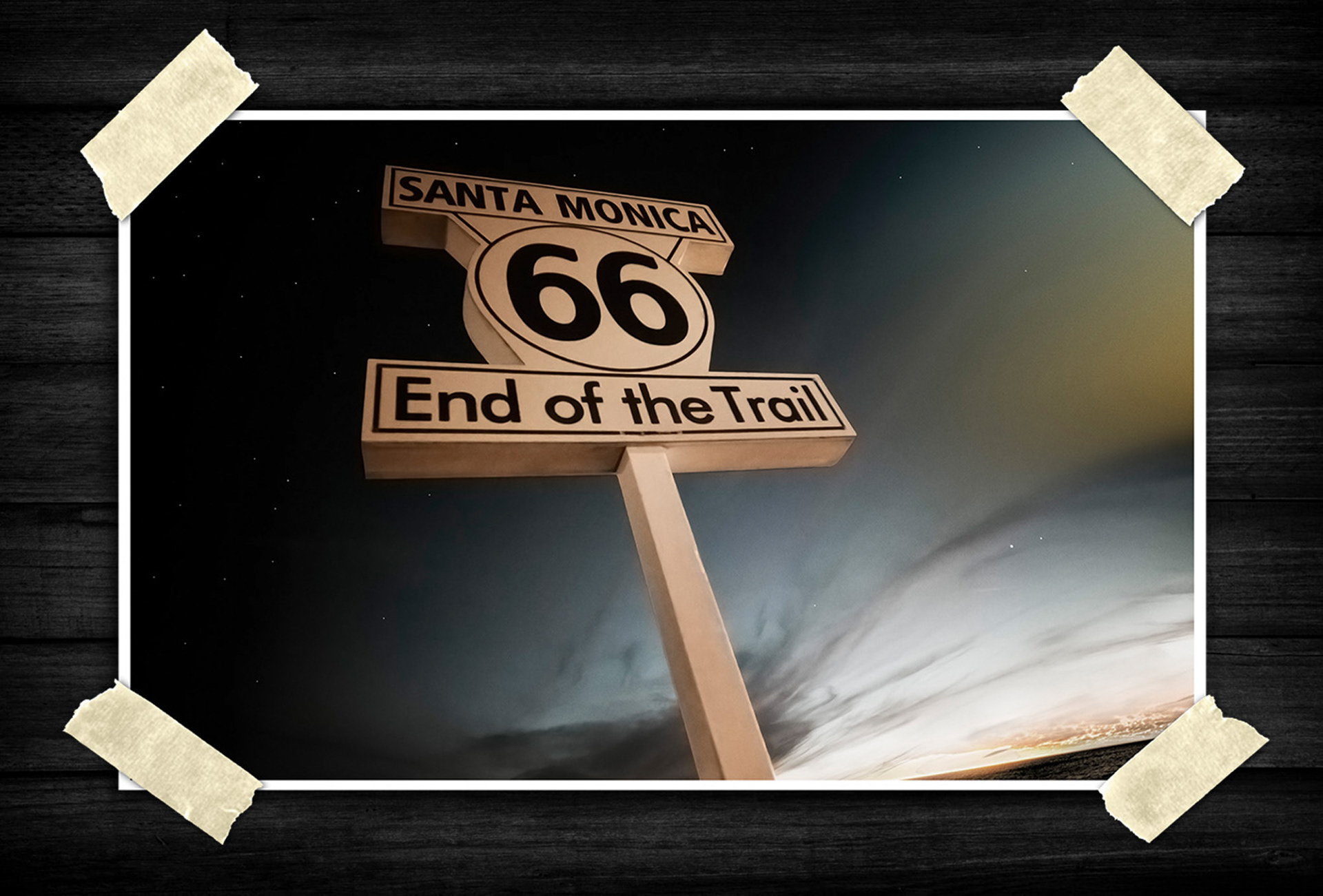 This image shows a stylized roadside sign reading “Santa Monica 66 – End of the Trail.” It is photographed from a low angle, making the sign appear tall and dominant against a dramatic night sky. The background has a dark, cinematic look with stars or bright points visible and streaked clouds near the horizon.
