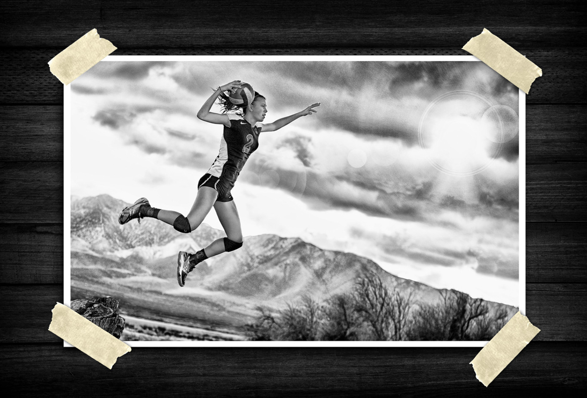 Black-and-white action shot of a volleyball player leaping into the air with a ball in hand, set against a dramatic mountain landscape and cloudy sky.