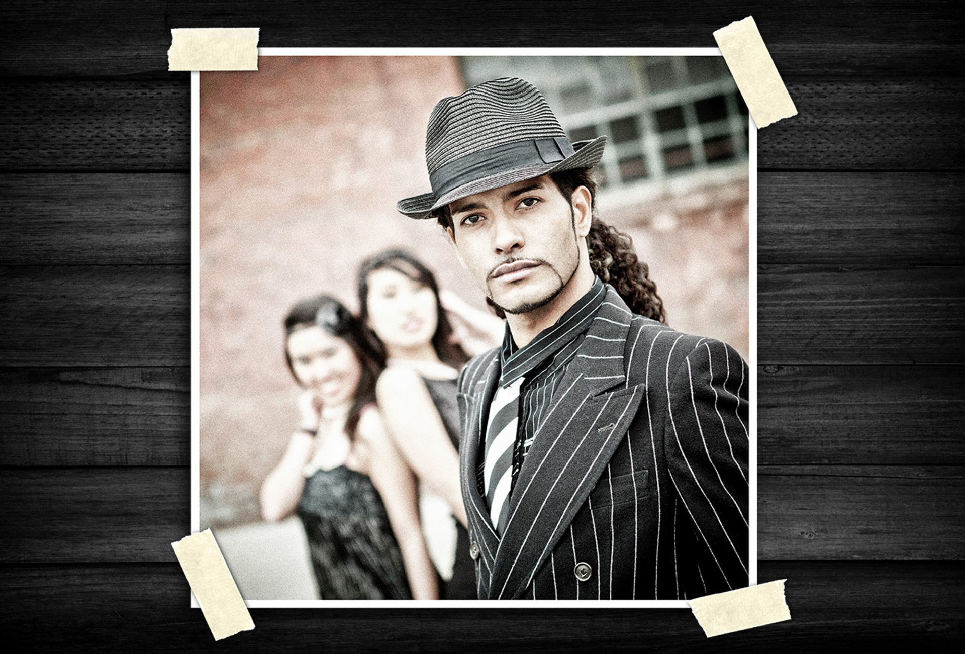 Fashion portrait of a man wearing a pinstripe suit and fedora hat, posed confidently in the foreground with blurred figures in the background.