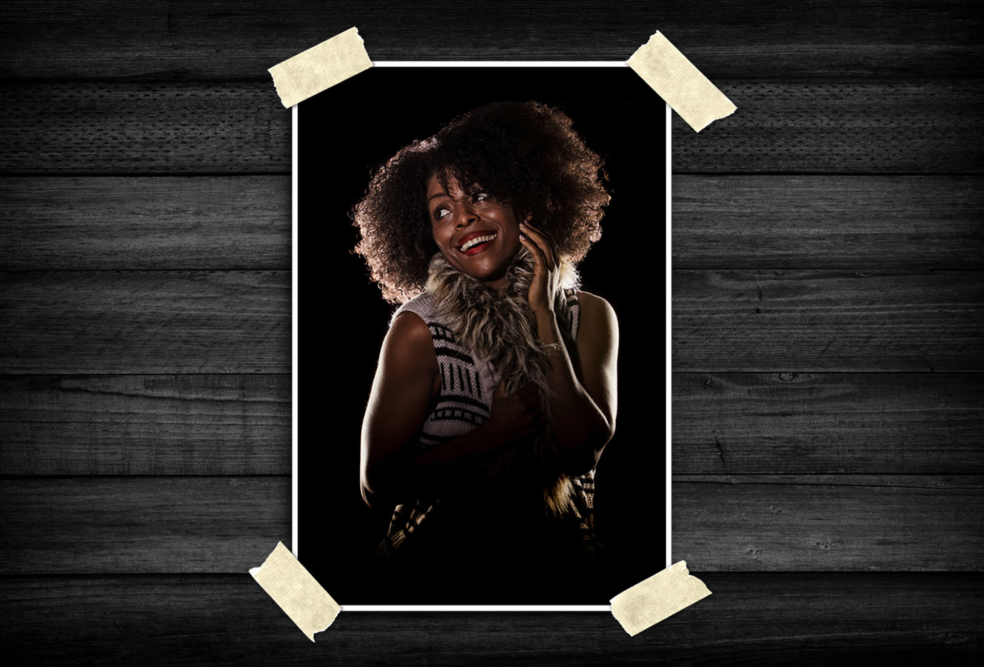 Portrait of a smiling woman with natural curly hair and textured fur-accented clothing, lit with dramatic studio lighting against a black background.