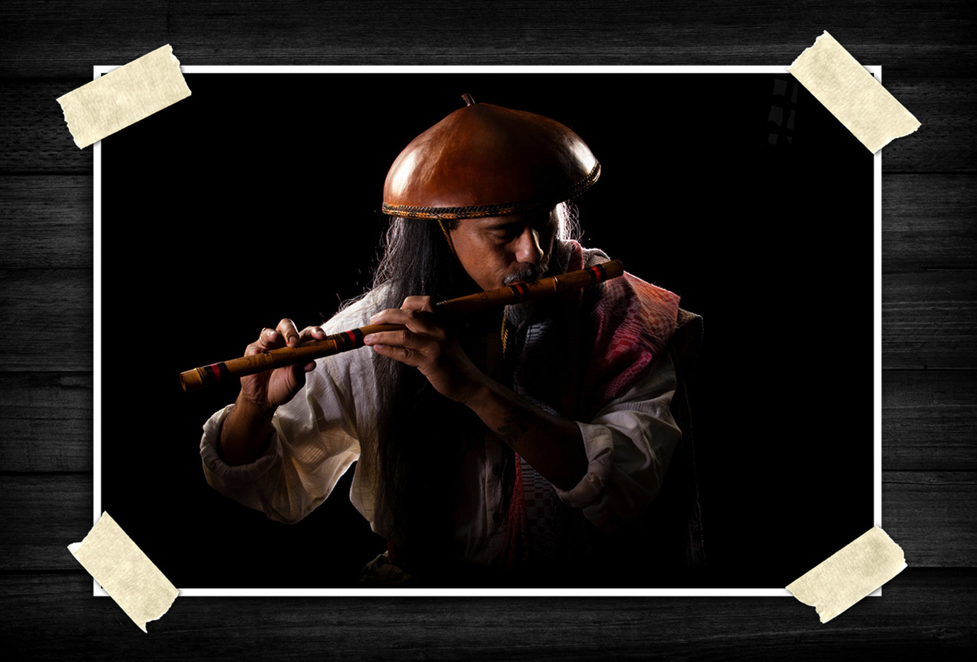 Portrait of a man in traditional attire playing a wooden flute against a dark background, illuminated with dramatic studio lighting.