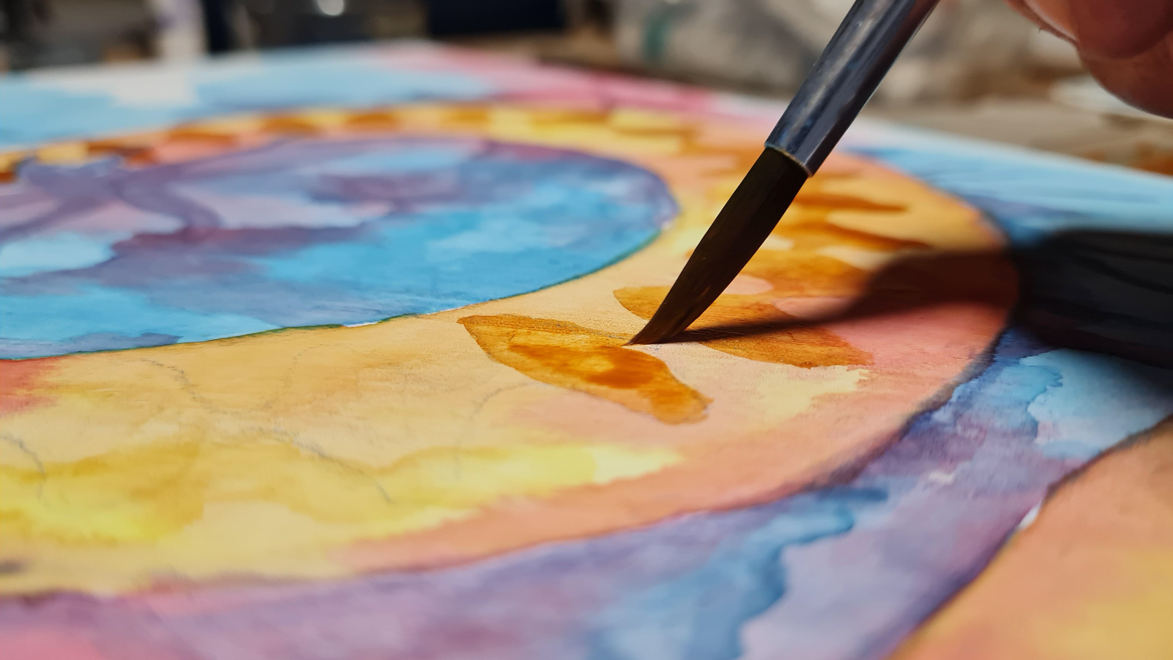 A detail of Lukasz Iskierka, Bristol-based artist, painting in his unique style, blending pencils and water colours. The image captures the tip of a brush touching the paper covered with a mixture of yellow and orange against a blue and purple background.