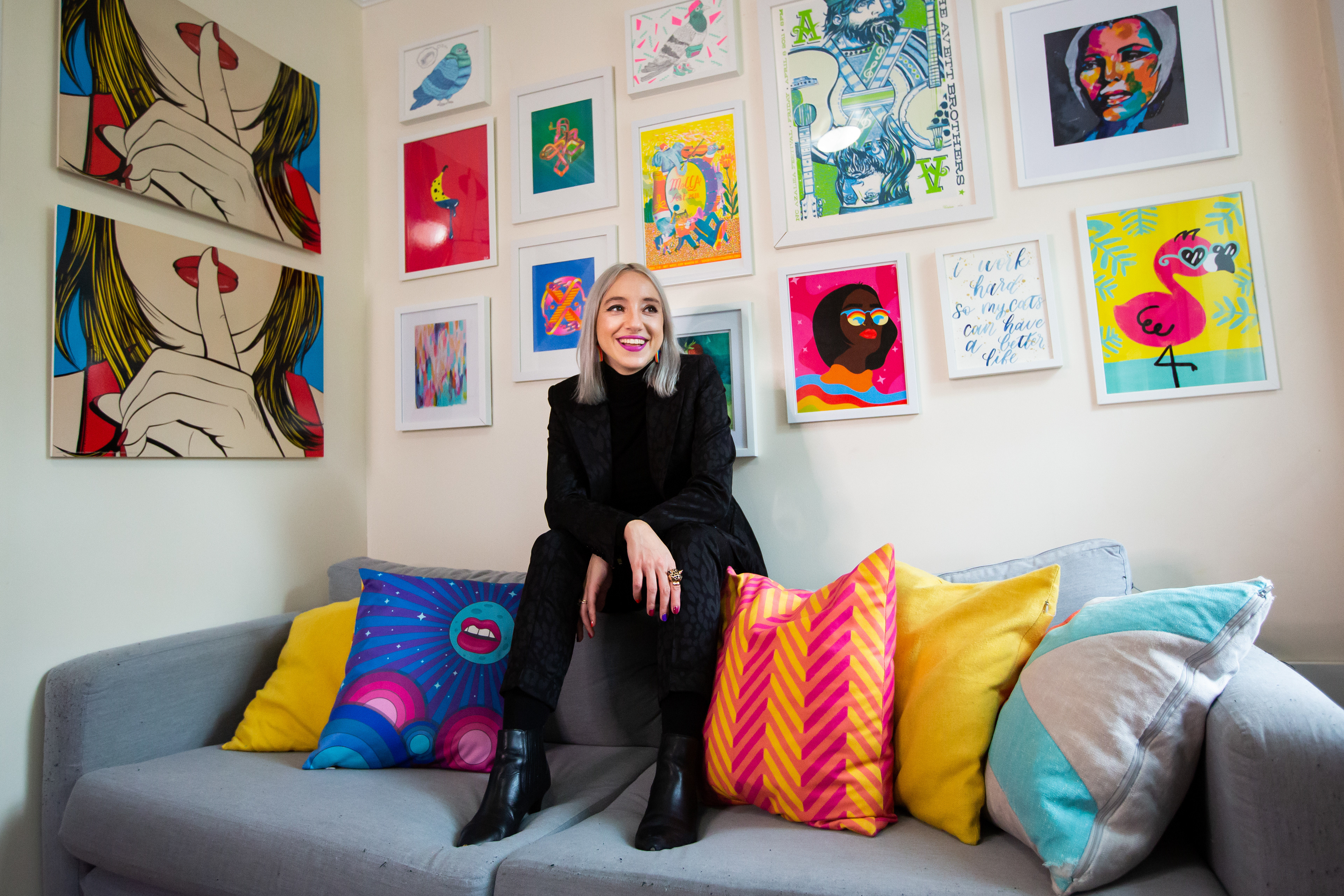 Barbara Astrini sits on a gray couch, in front of a wall gallery of colorful paintings
