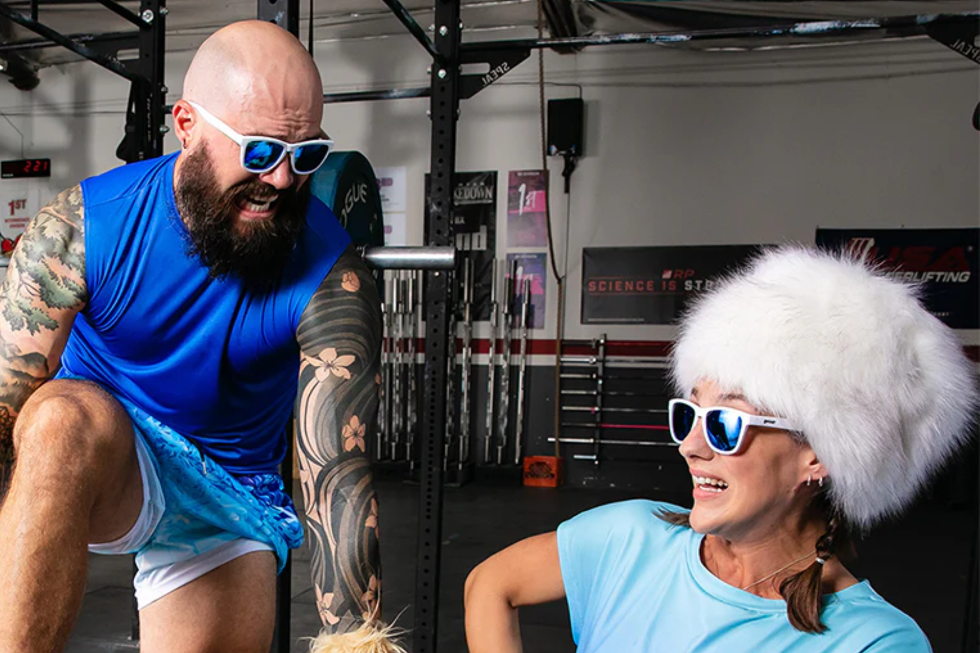 A man and a woman in a gym wearing goodr sunglasses with white frames and mirrored blue lenses