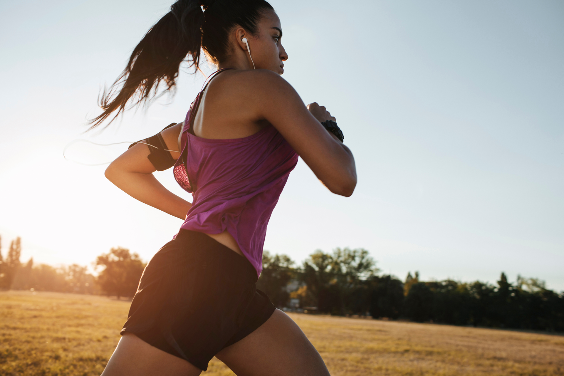 A brunette woman with a ponytail is running outside on a clear day with trees and a grassy field in the background. She is wearing a purple racerback tank top, black shorts, has white earbuds in, and a black phone armband on her left arm.