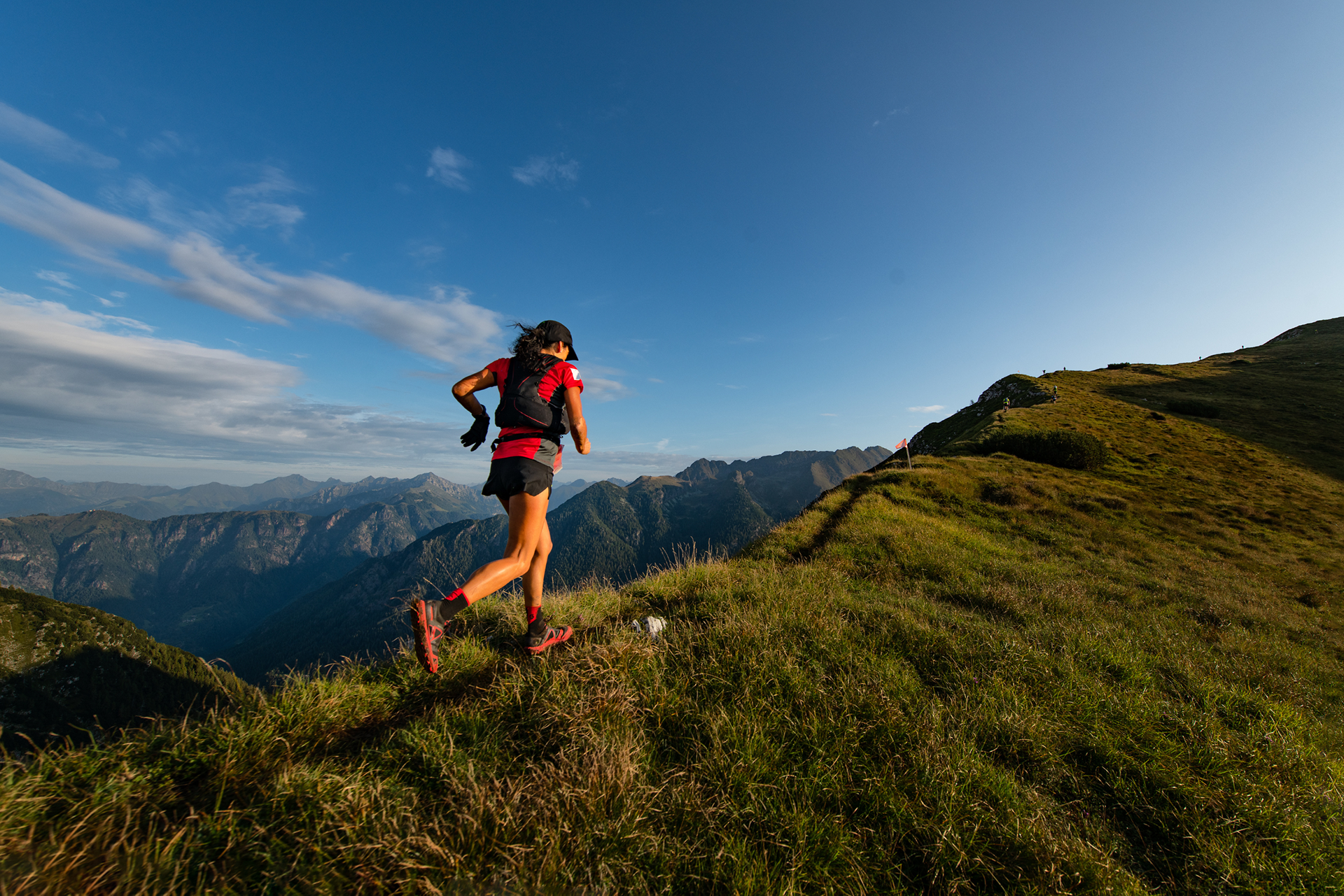 A woman with dark hair is running atop a lush green mountain trail with multiple mountain ranges in the background. She is wearing a red and black t-shirt, socks, and shoes, and black shorts and cap. She also has on a red and black running backpack.