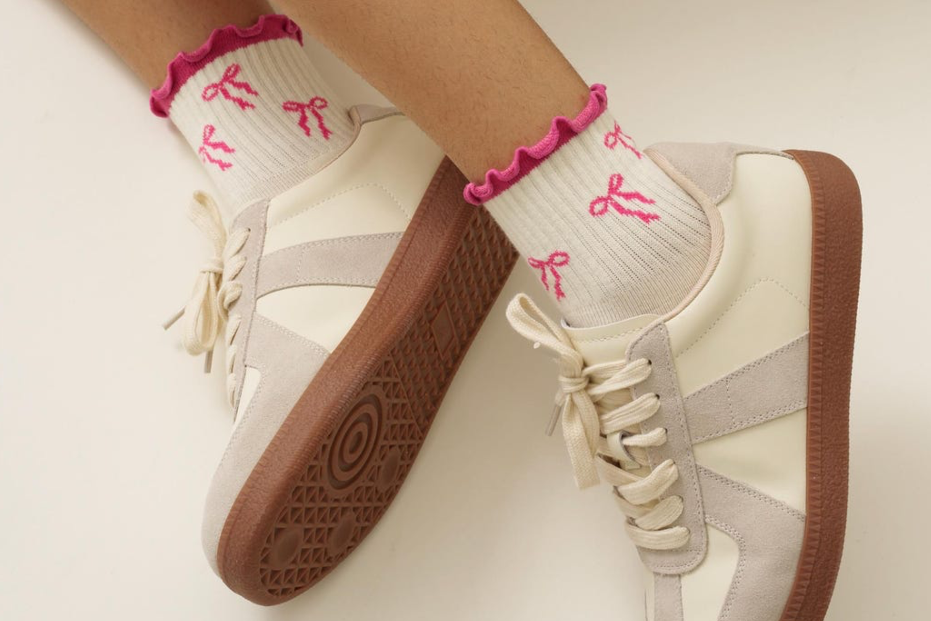 A pair of legs with white Shapes Studio half calf ruffle socks with a pink trim and bows and white and grey sneakers on