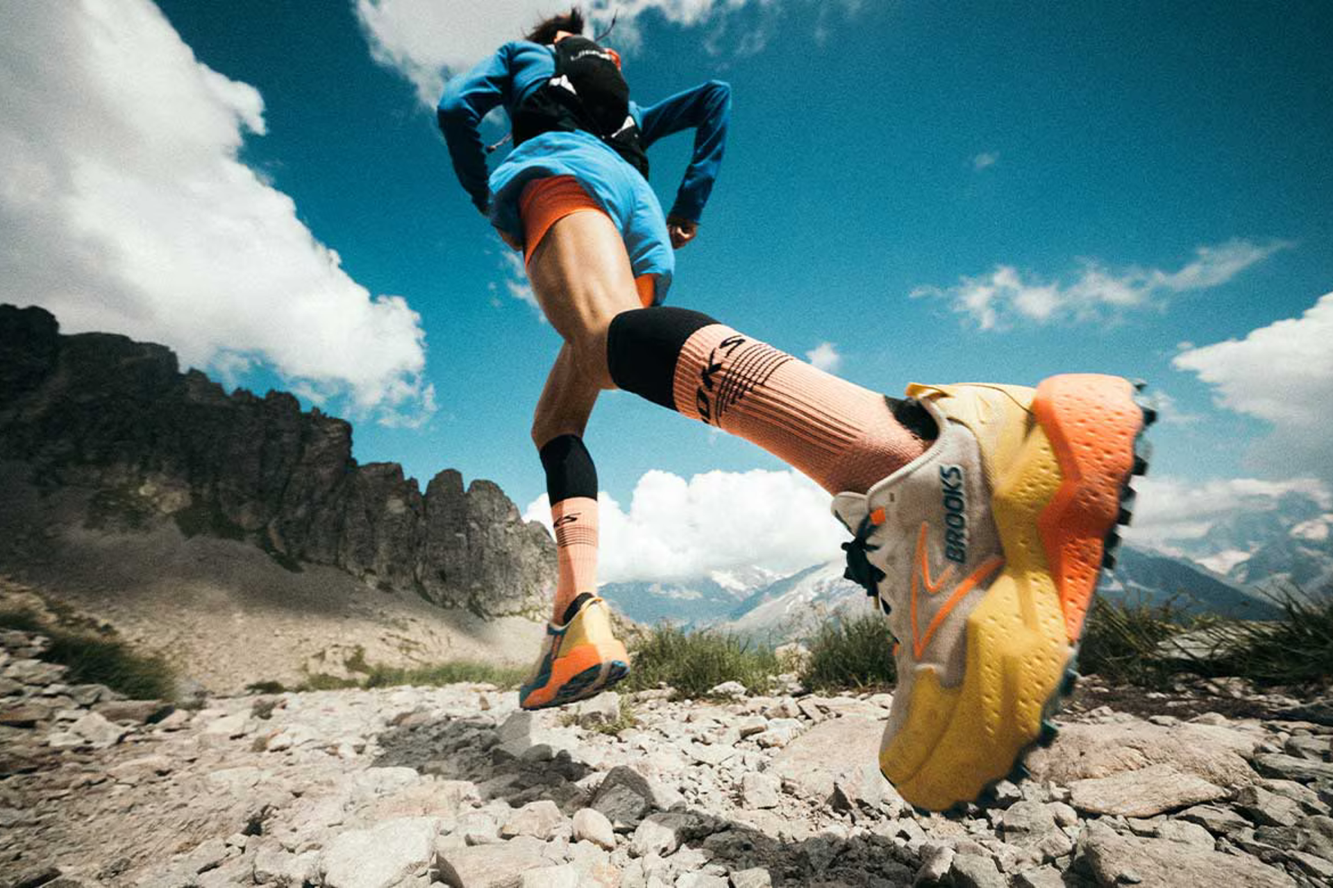 Runner on a rocky mountain trail wearing white Brooks trail running shoes with yellow and orange soles, orange and black socks, bright blue long sleeve and shorts, orange shorts underneath, and a black running backpack.