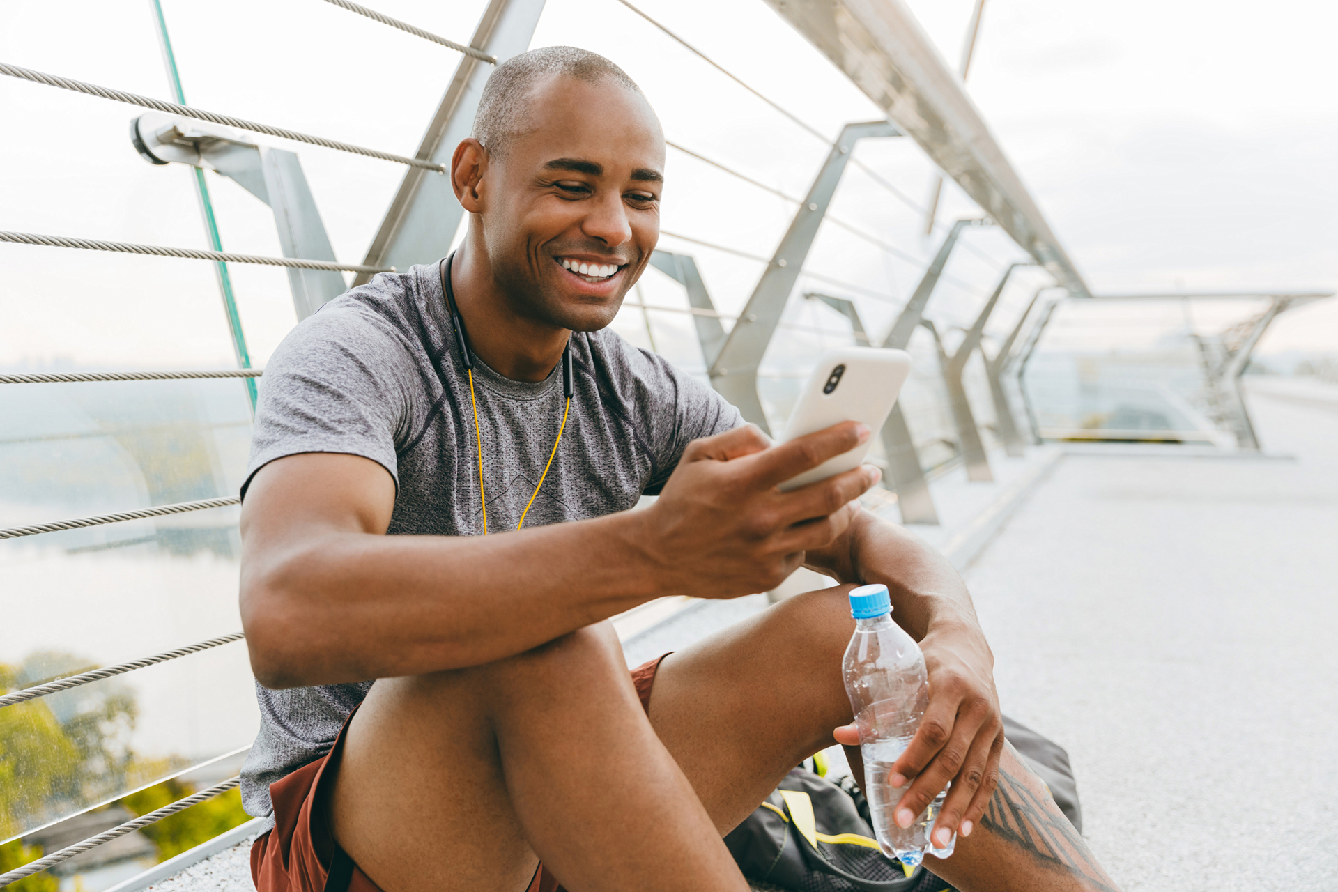 A man sitting outside on a pedestrian bridge wearing a gray t-shirt and sepia shorts is smiling while looking at his white phone in his right hand. He is holding a half empty plastic water bottle with his left hand.