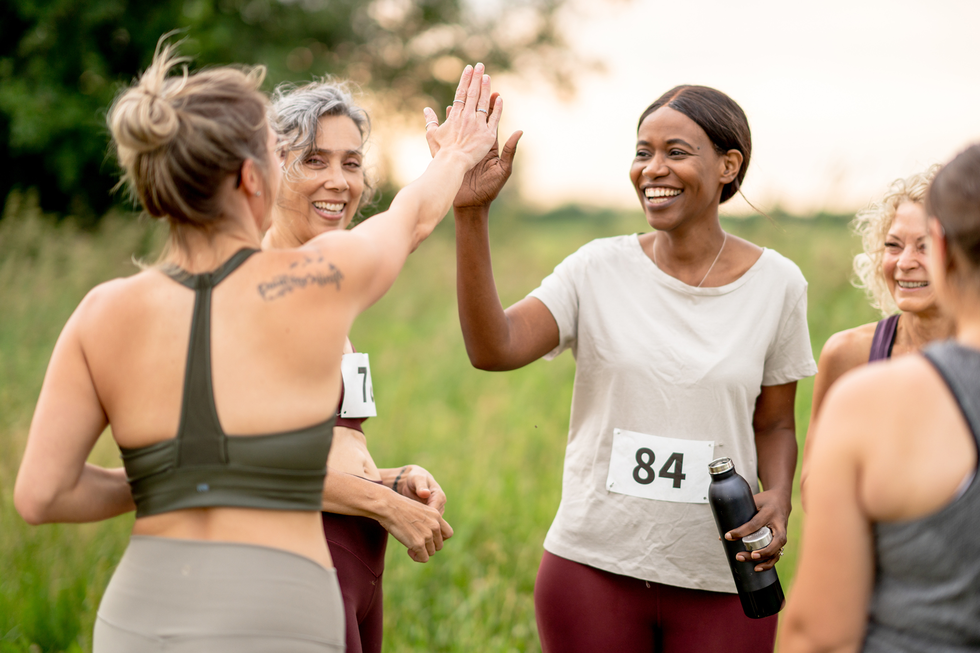 A diverse group of five women smiling while outdoors surrounded by a grassy field and trees. They are wearing exercise clothes and have race bibs on. Two are high fiving each other.