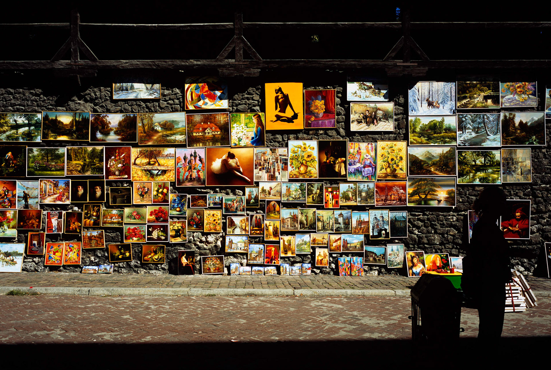 Paintings for sale hung on historic city walls in Krakow, Poland, with woman in shadow in foreground
