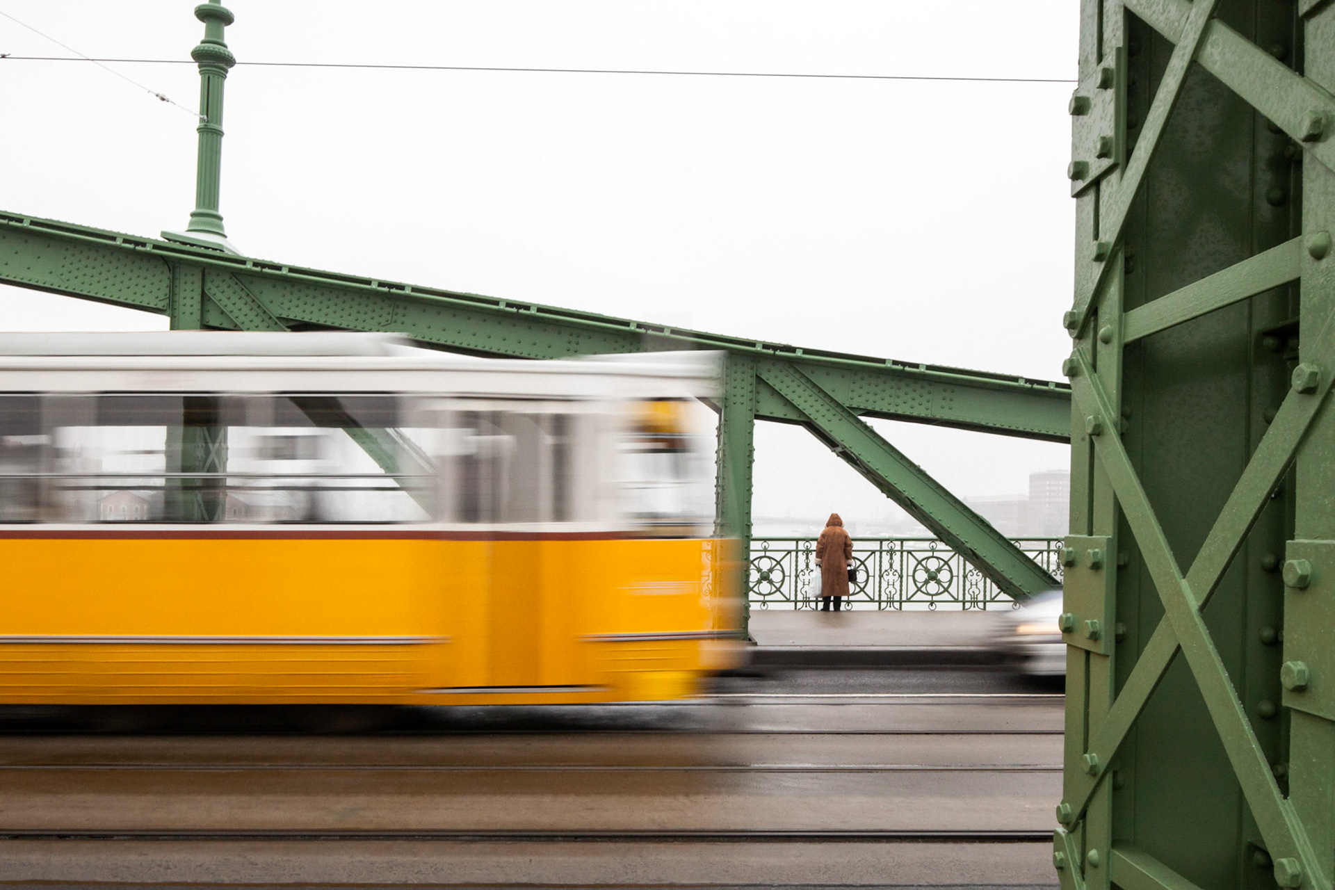 Lady wearing winter coat standing on Liberty Bridge with back turned looking at Danube River as yellow and white tram passes in the foreground, Budapest, Hungary