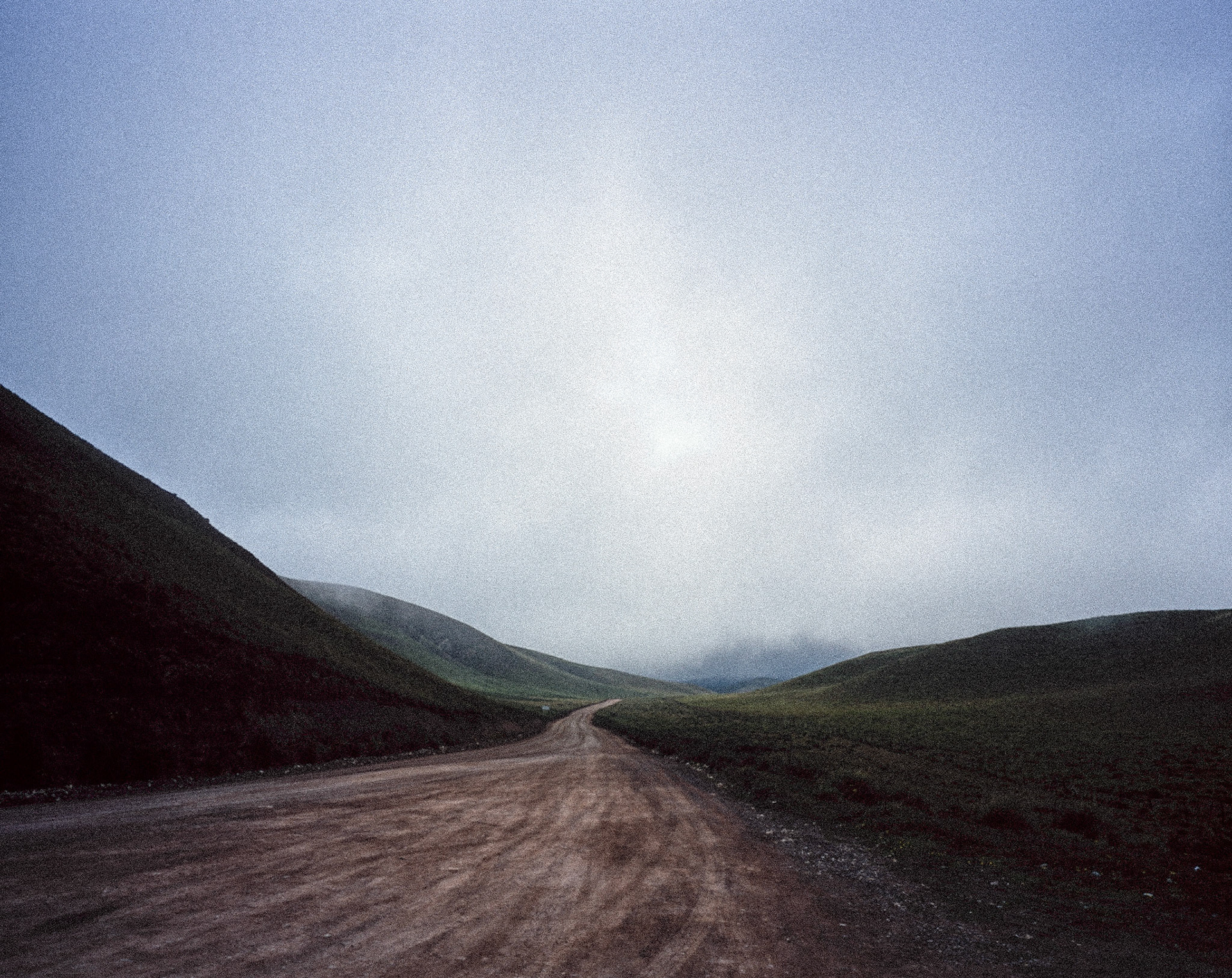 Slightly winding dirt road surrounded by green hills heading through clouds in distance to Altiplano, Bolivia