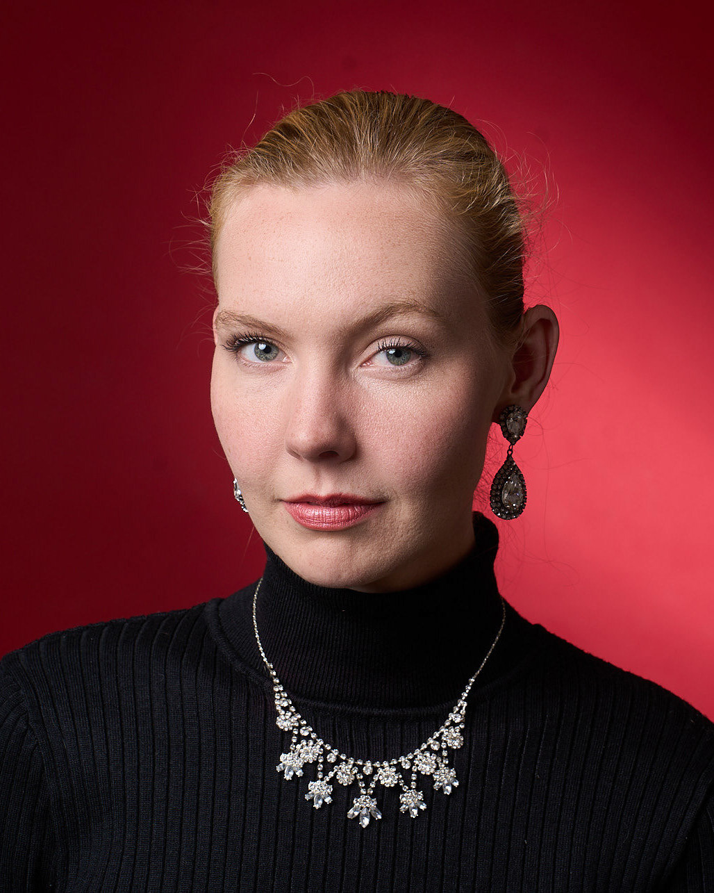 A portrait headshot of a person with blond hair in a black turtleneck, wearing a matching necklace and earrings, poses against a plain red background.