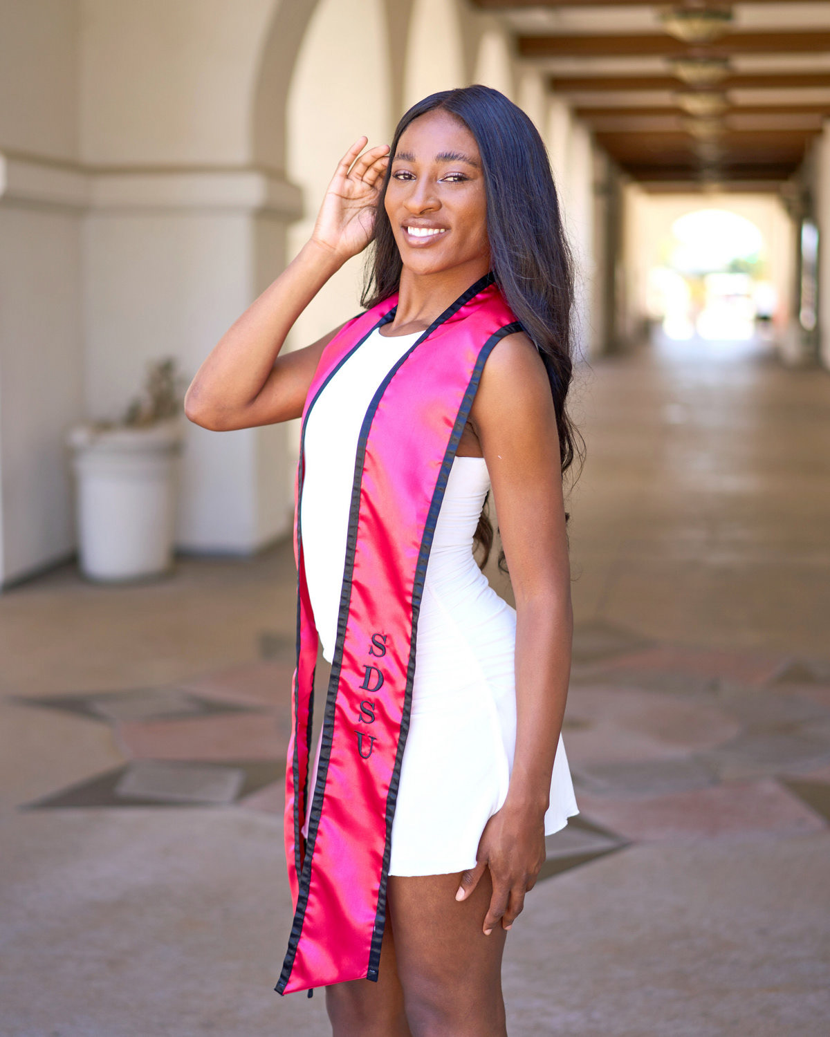 Smiling graduate wearing a white dress and red stole standing in an arched hallway