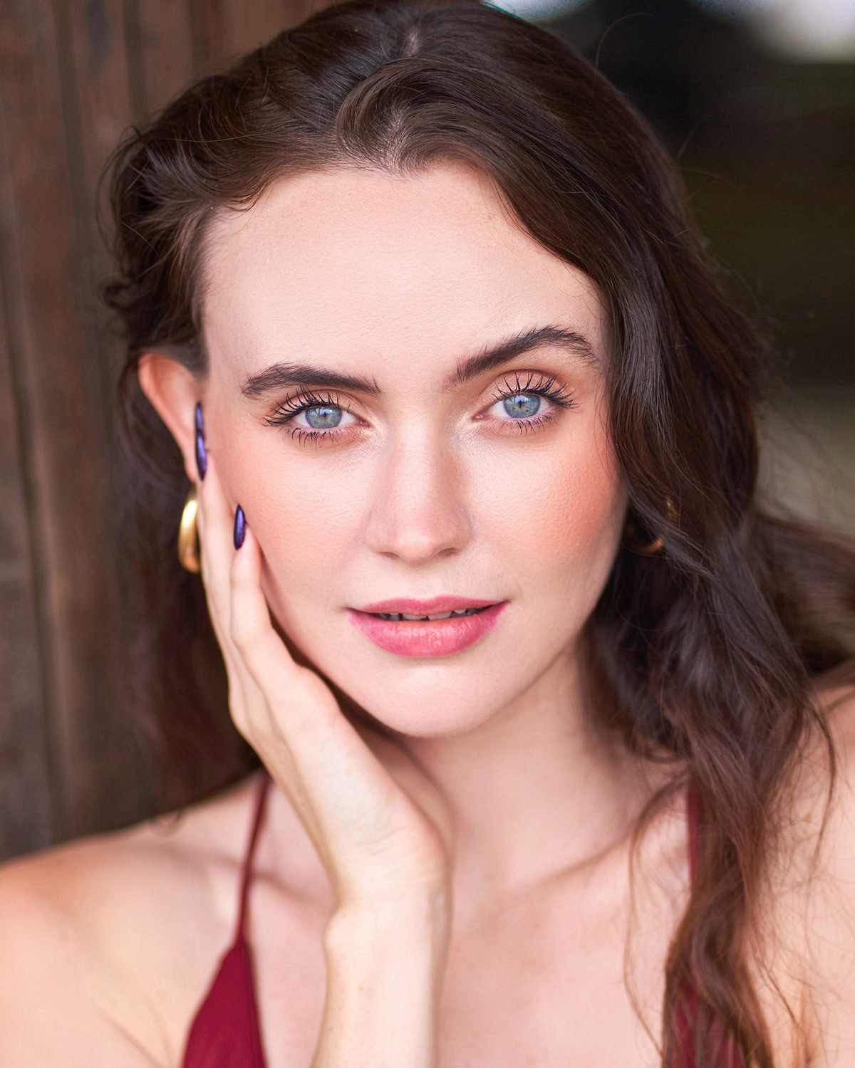 Close-up portrait of a woman with blue eyes, long wavy brown hair, and natural makeup, posing with one hand touching her face, wearing gold hoop earrings and a burgundy top