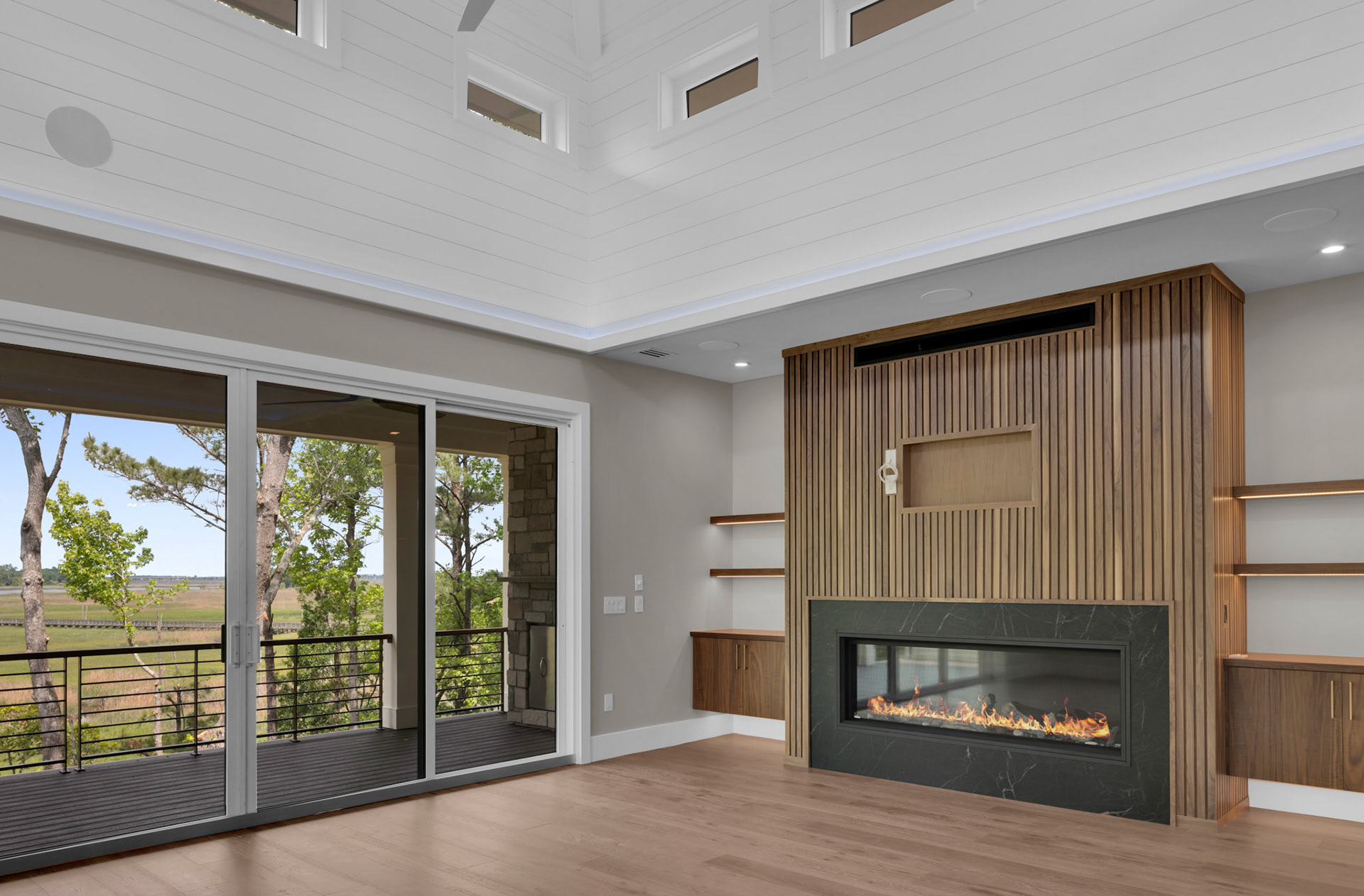 Custom linear fireplace wall with vertical wood slat detailing, floating built-ins, and full-height glass doors bringing natural light into this luxury coastal home.