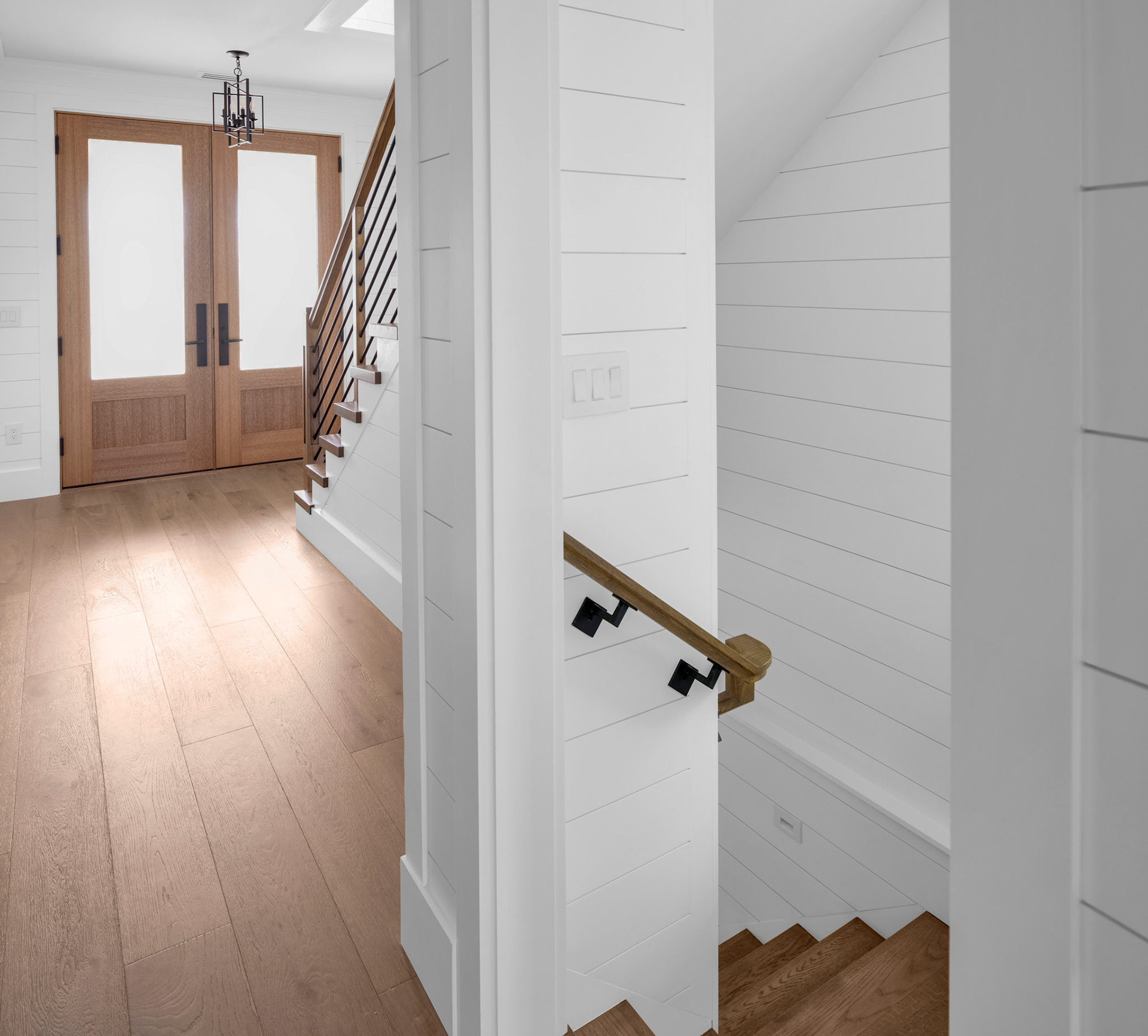 Bright interior hallway featuring modern coastal finishes, wide-plank flooring, custom staircase railings, and natural light from oversized entry doors.