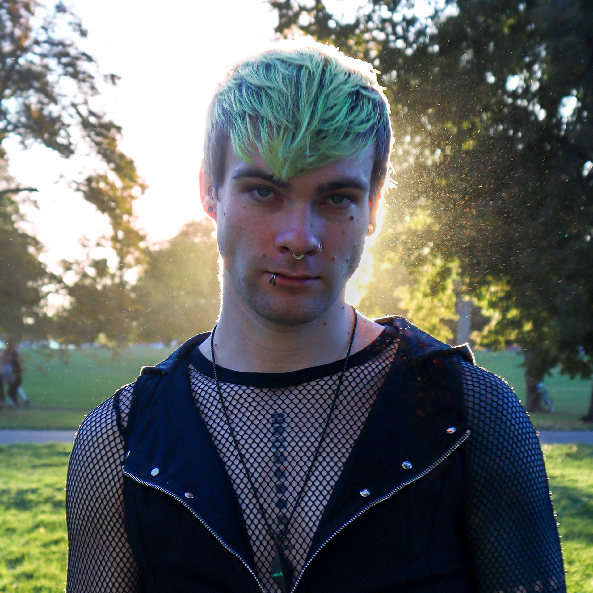 A fabuslously attractive and professional individual smolders at the camera, a slight, mischievous smile implied in their eyes. The setting sun frames their face with dust motes and lens grit, and there are lush trees and grass behind them. Their outfit is a black mesh shirt layered under a black zippered vest. A long chain necklace with a single understated pendant hangs around their neck and frames a series of circles and rectangles on their chest: a tattoo.