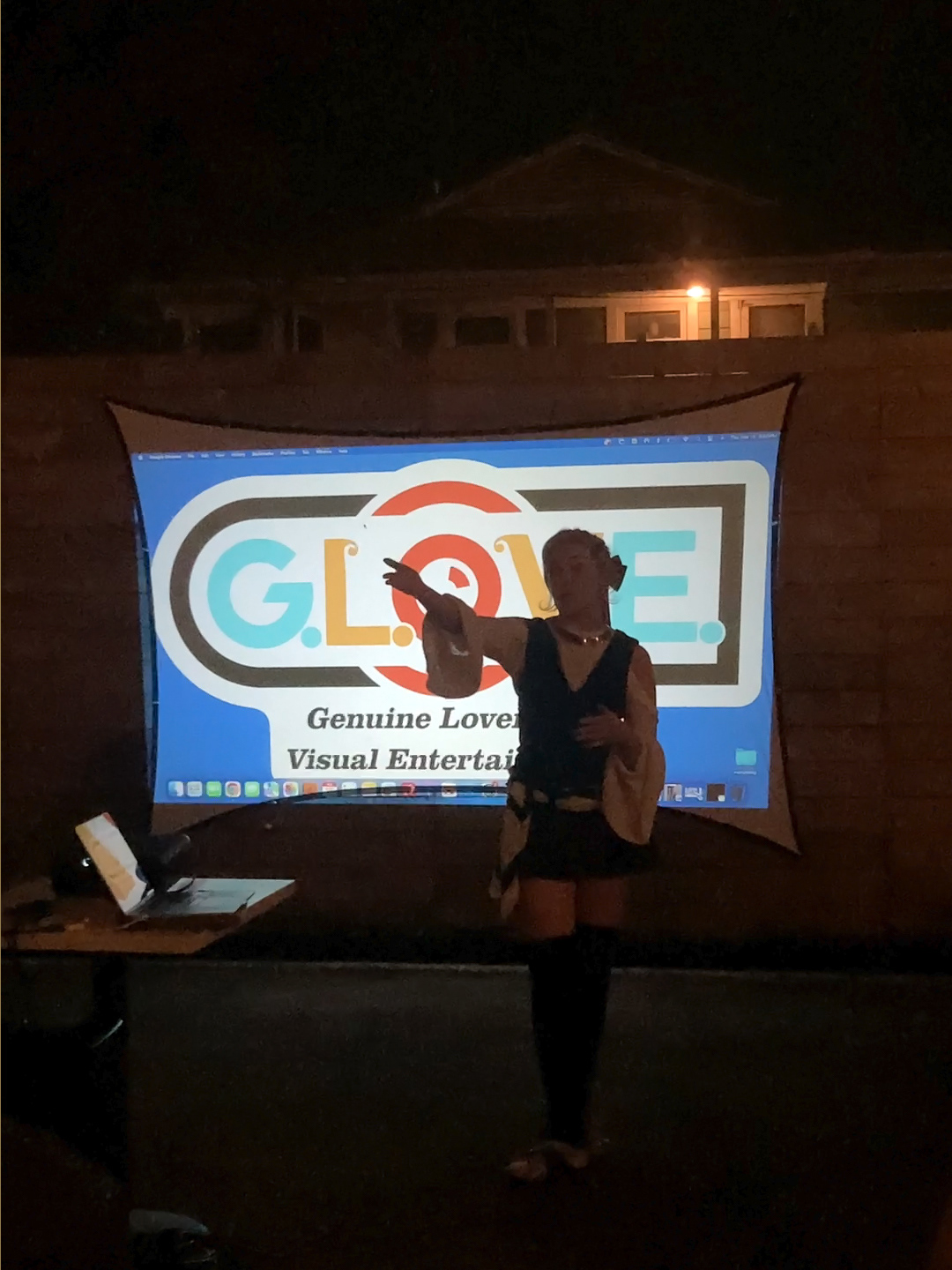 Chloe, the film showcase's host and founder, stands gesturing in front of the GLOVE logo, projected on a screen, outdoors at night.
