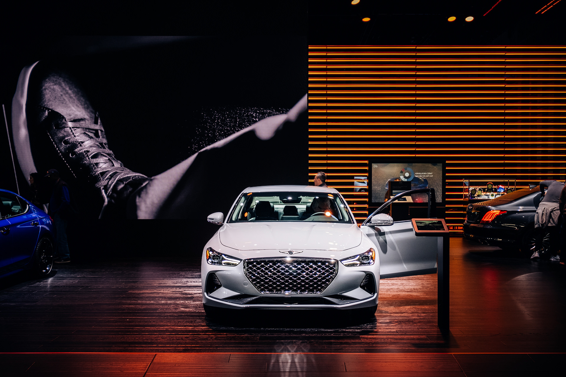 White Genesis sedan displayed beneath exhibition lighting at an indoor auto show.