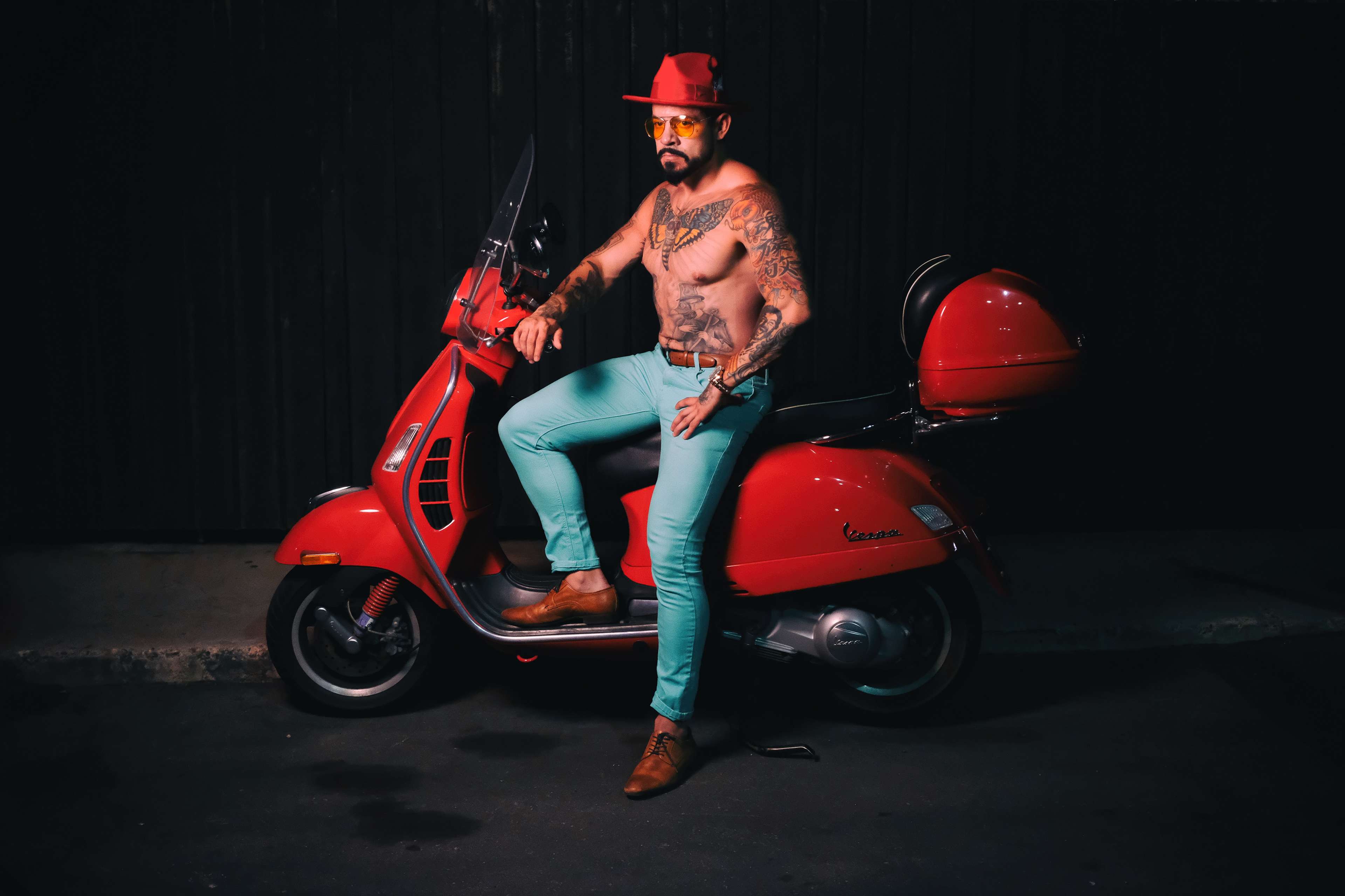 Fashion portrait of a tattooed man wearing a red hat, tinted glasses and turquoise trousers while seated on a red scooter.