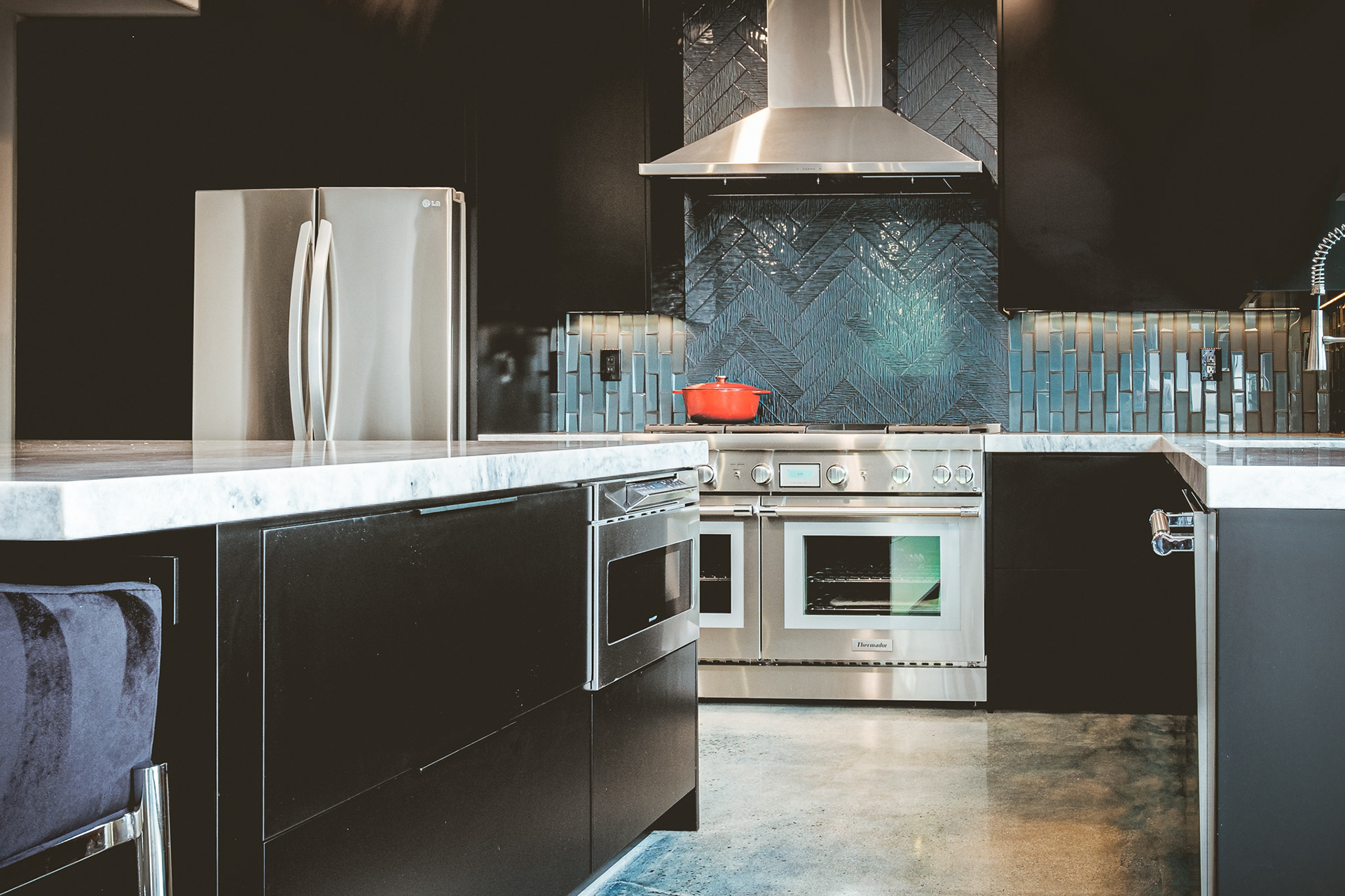 Modern kitchen with dark cabinets, a marble island, stainless steel appliances and a blue tile backsplash.