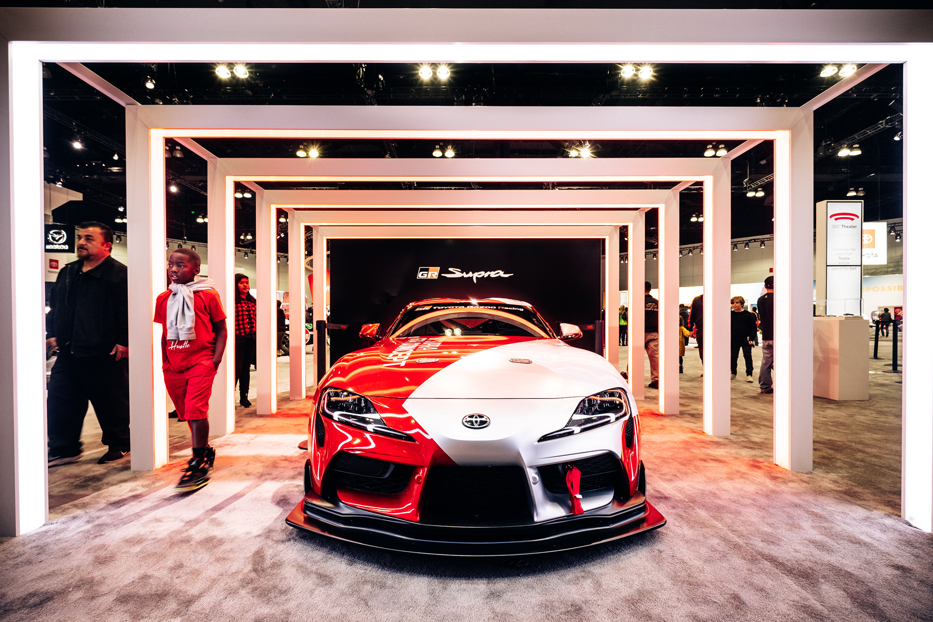 Red and white Toyota GR Supra beneath illuminated display frames at an auto show.