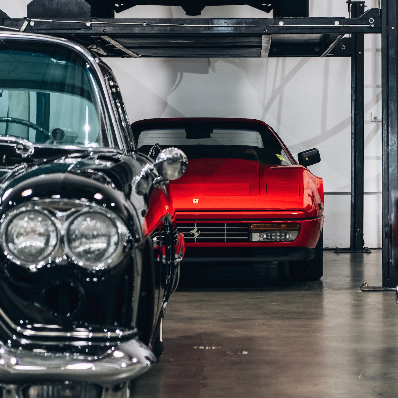 Black classic car in the foreground with a red Ferrari parked behind it inside a garage.