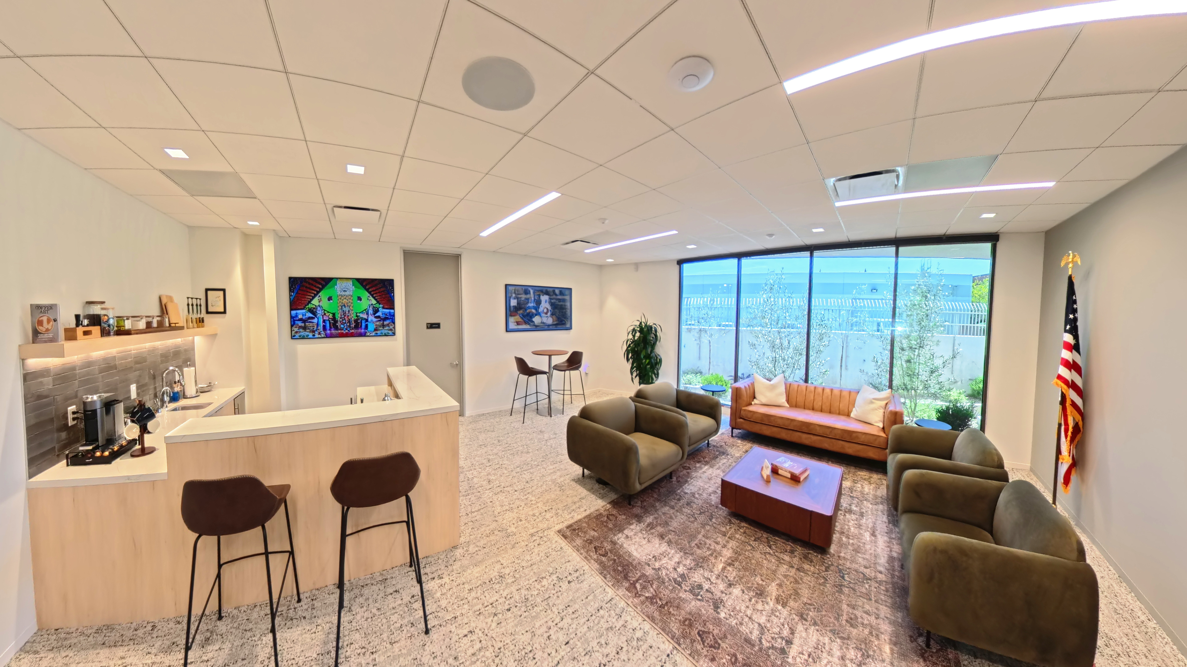 Office lounge with a kitchenette, counter seating, green armchairs and a sofa beside large windows.
