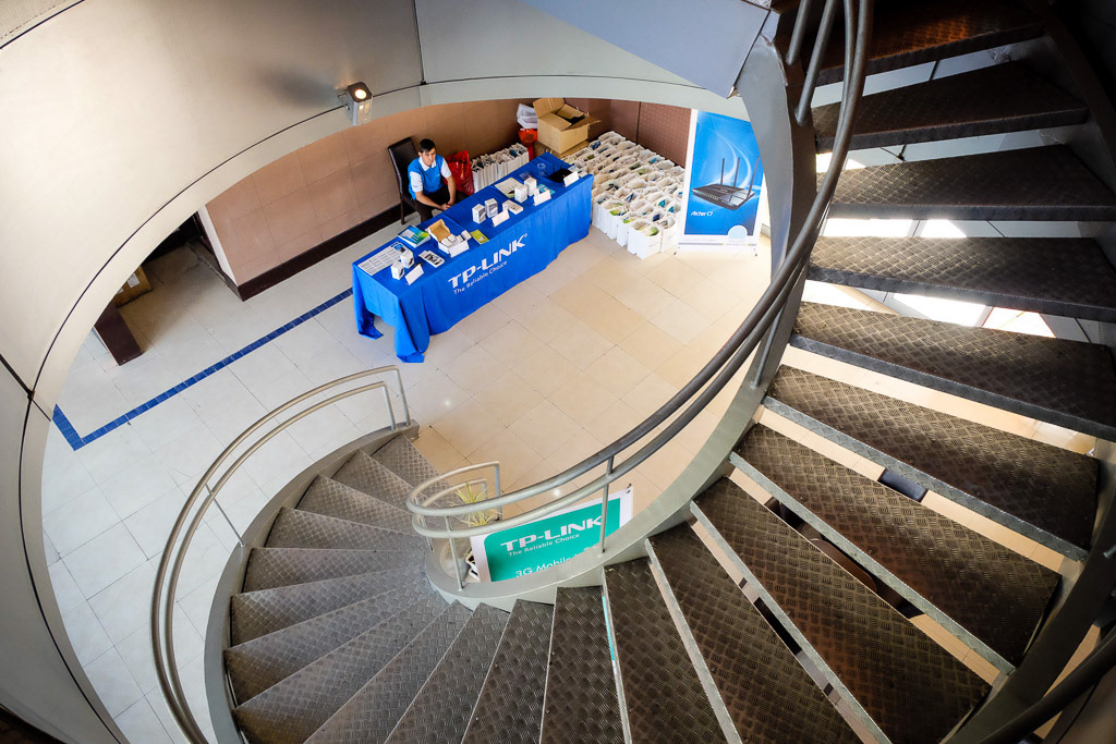 View down a curved staircase toward a TP-Link event display and welcome desk.
