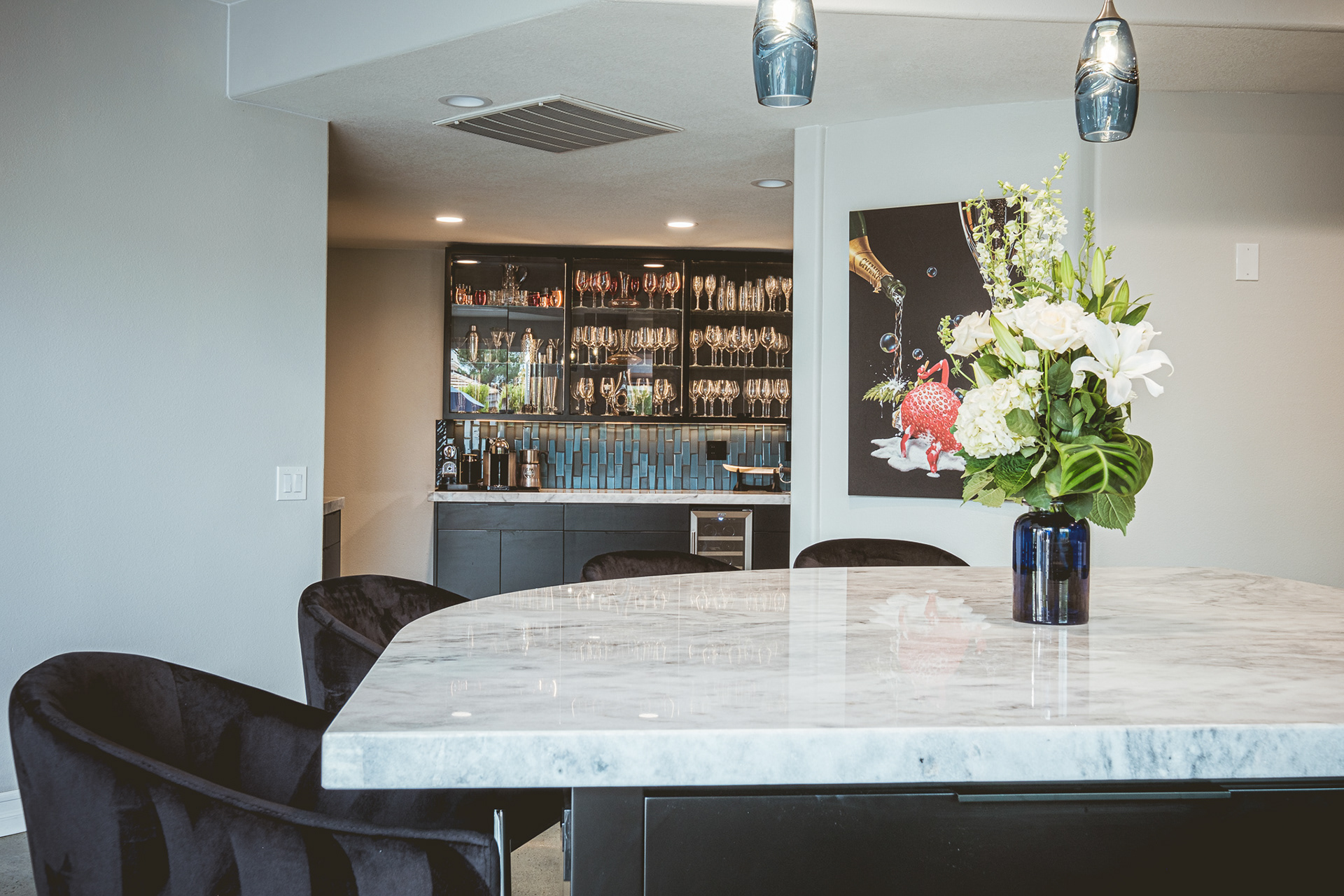 Marble kitchen island with dark stools, pendant lights and flowers, looking toward a home bar.