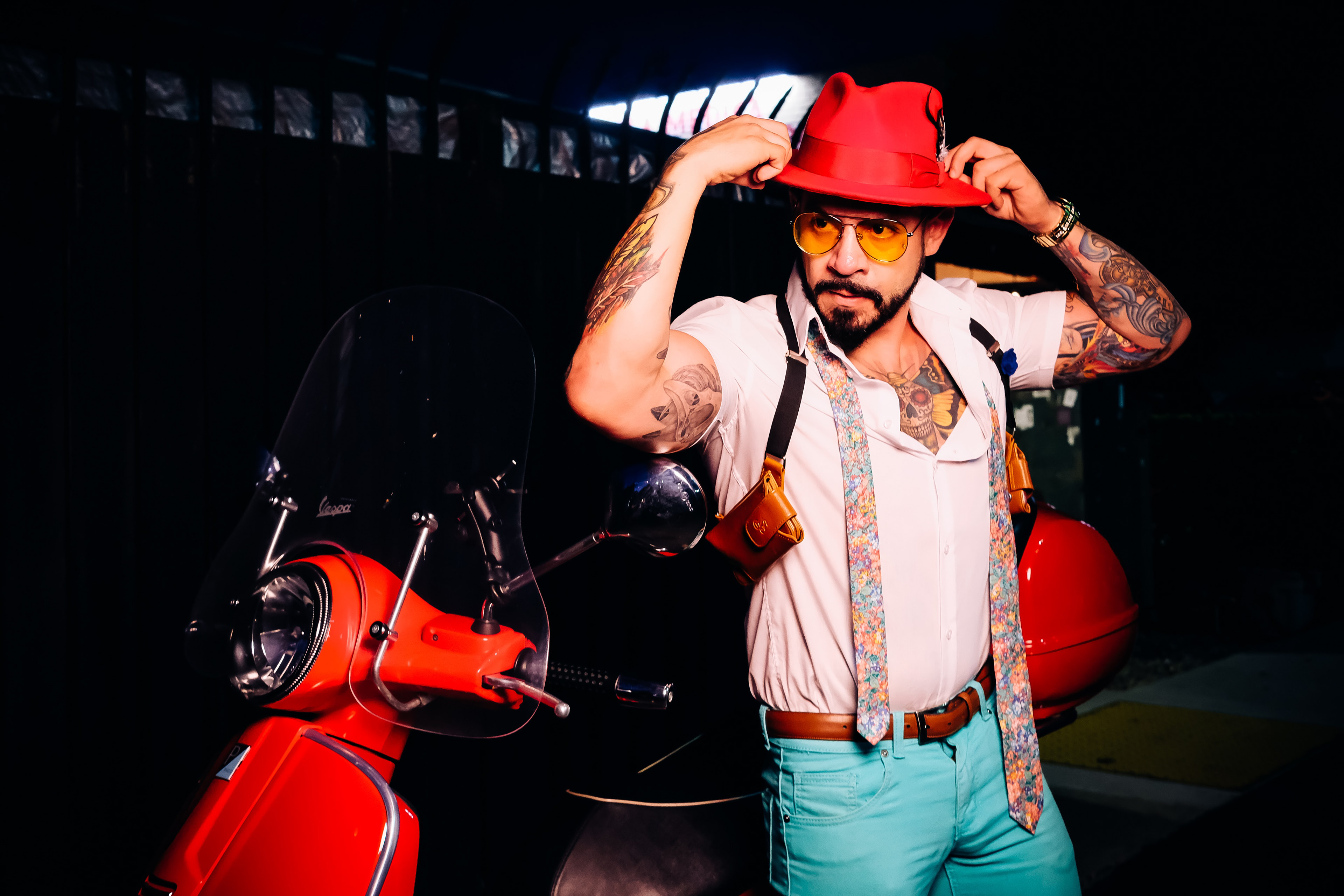 Fashion portrait of a man adjusting a red hat beside a red scooter, wearing suspenders and turquoise trousers.