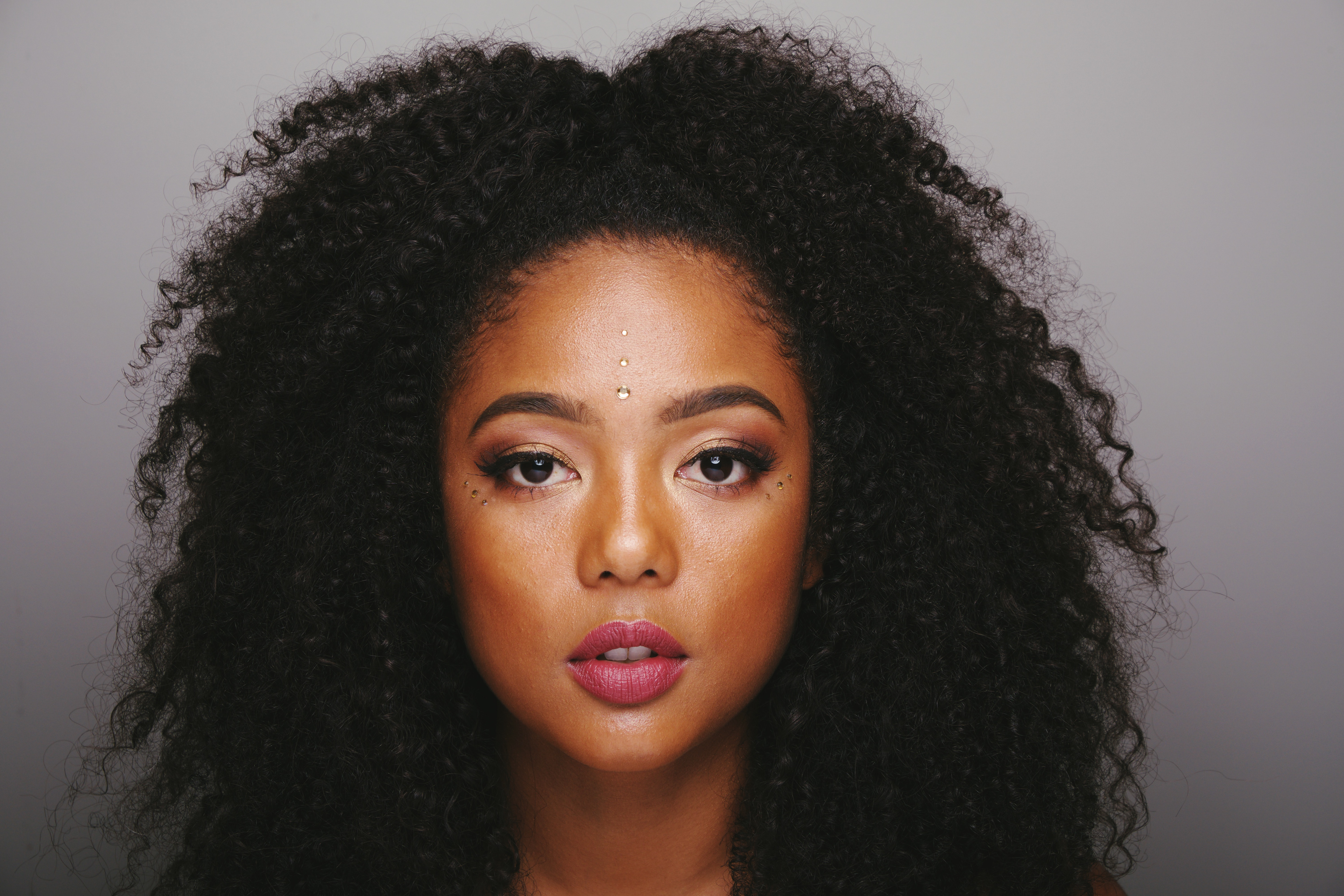 Close-up studio portrait of a woman with voluminous curly hair, decorative face gems and magenta lipstick against a gray background.