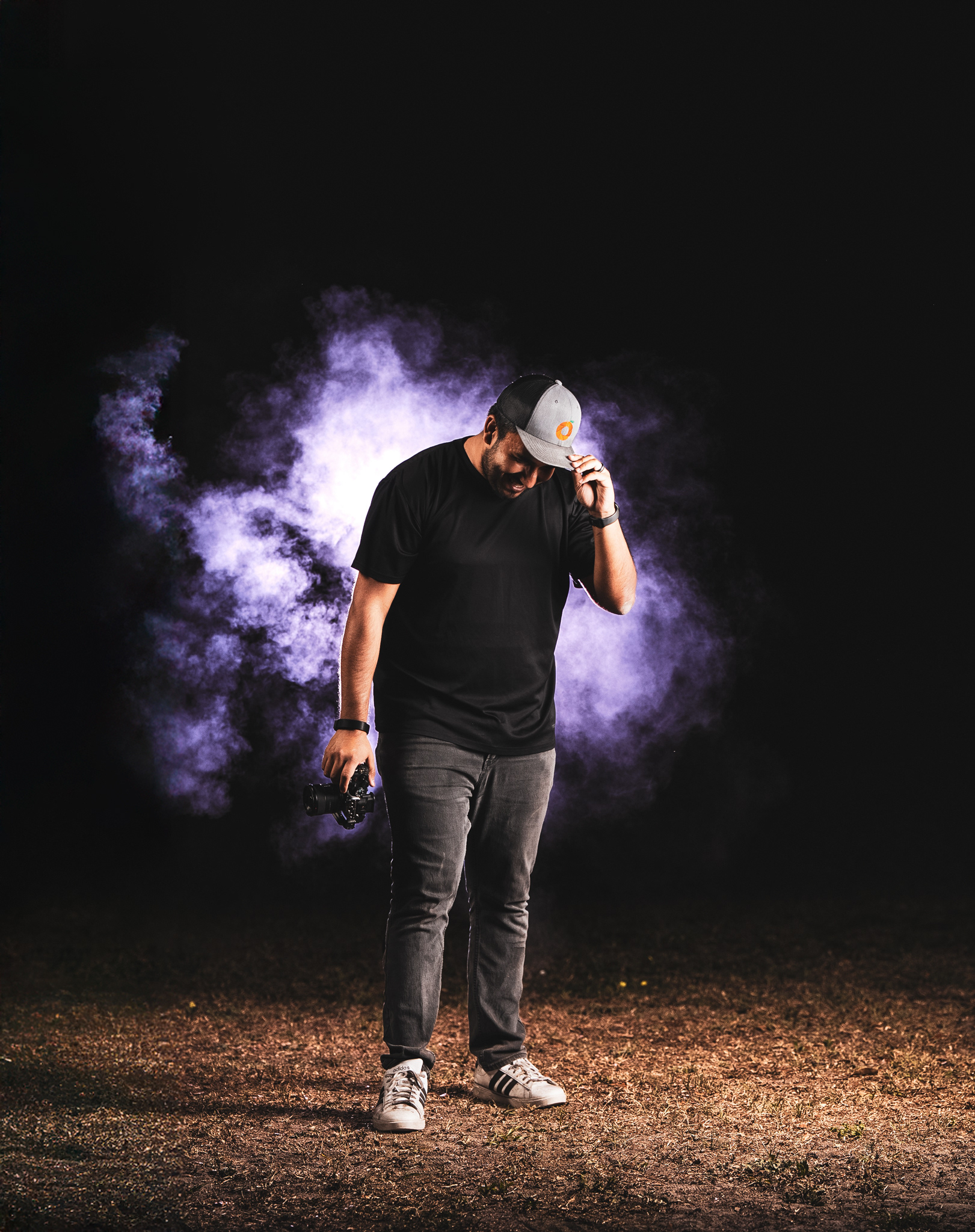 A creative portrait of photographer Richie Hartig. He's standing in a dark, atmospheric setting, illuminated by purple gelled rim lighting and smoke. He is looking down, holding a camera in one hand and the brim of a trucker hat with the other, demonstrating a cinematic off-camera flash setup.