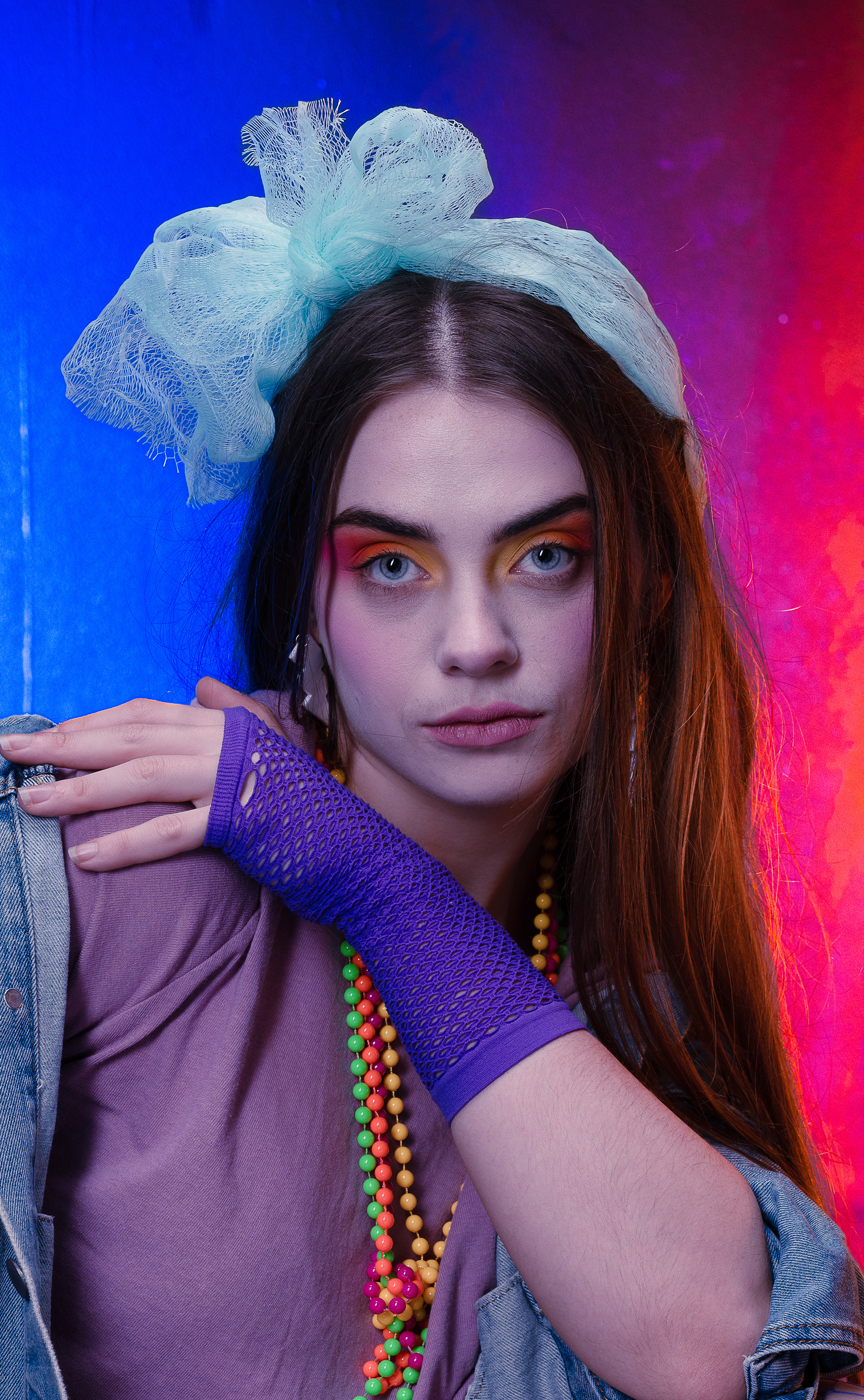 Material girl - Portfolio shoot - A young woman with long hair wears bright make-up and has a big bow in her hair, she wears bright beads around her neck and has a purple fishnet glove on her hand which is placed acros her body and rests on her right shoulder. She looks directly at the camera. 