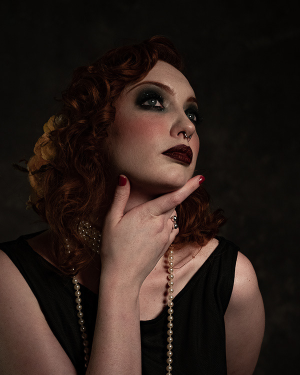 Roaring 20's shoot - a woman with dark eye make up hold her hand up to her neck she is gazing off into the distance 