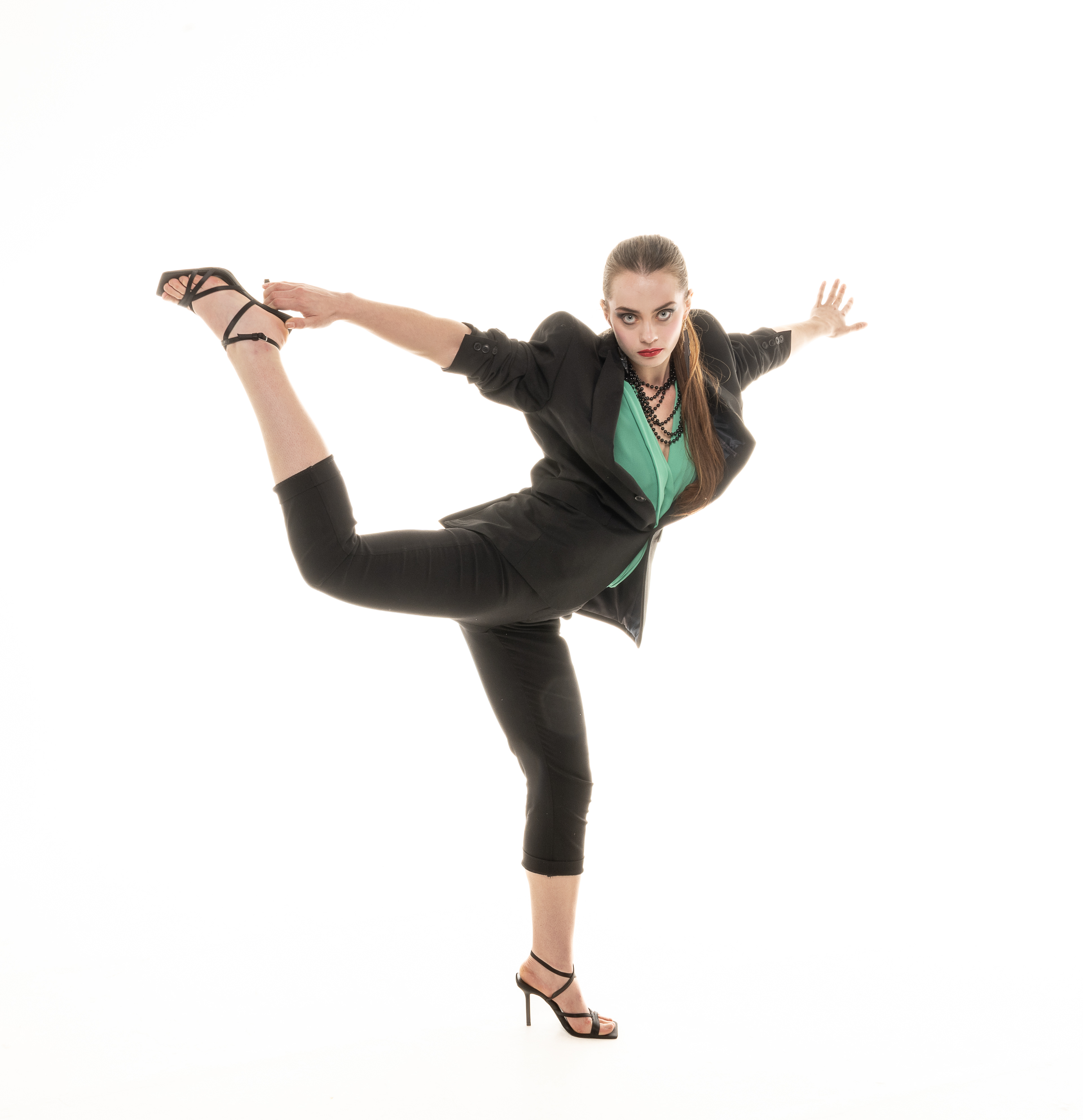 Eighties Vamp - Portfolio Shoot - A young woman wearing a black suit and high heels is stood on her left leg and has her right leg high behind her , she is looking straight at the camera