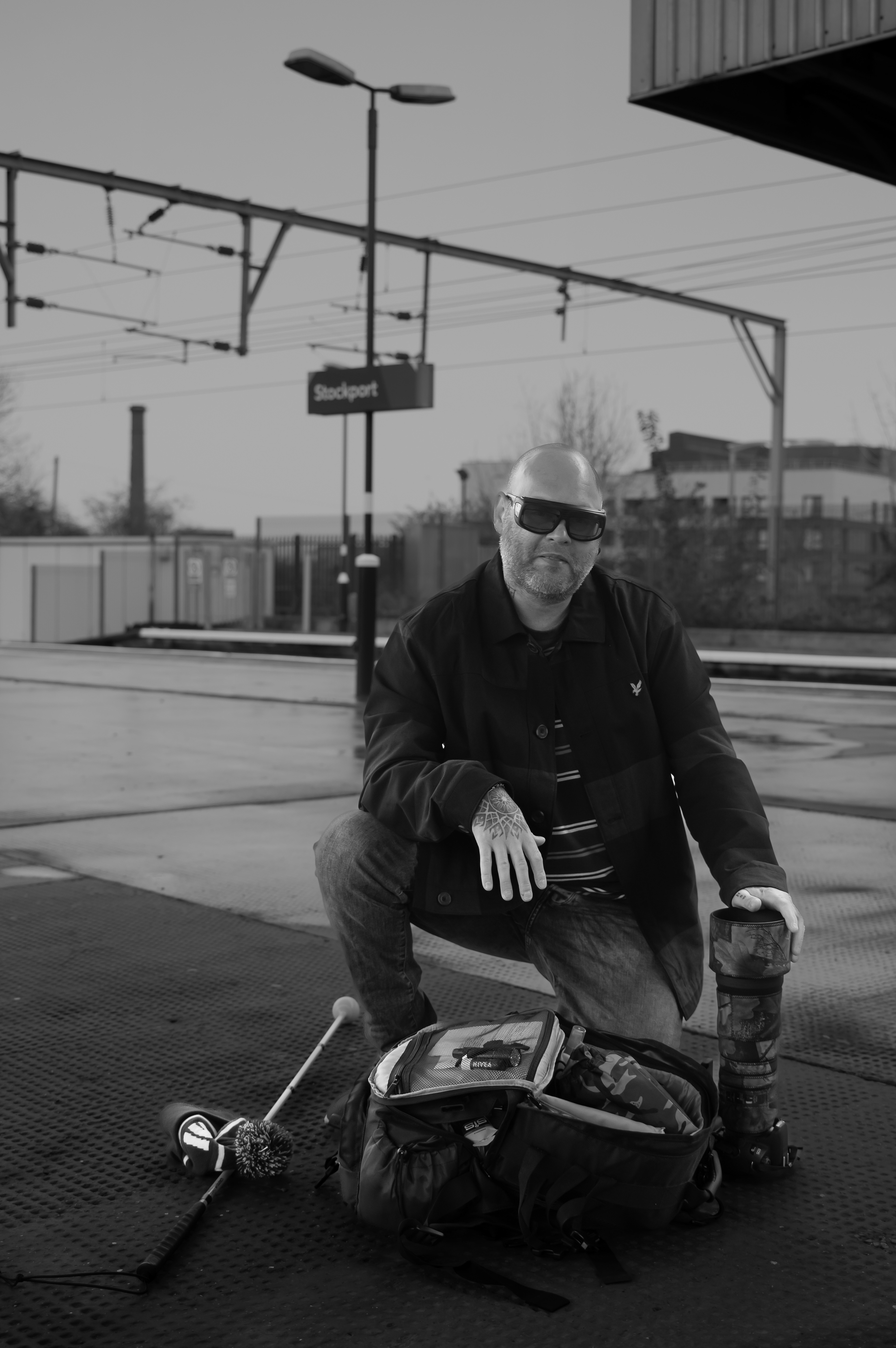 Women in Photography Exhibition 2024 -  a man kneels down on the platform of a train station. by his side is a white Caine, there is a bag in front of him and he has his one hand placed on a long lens  