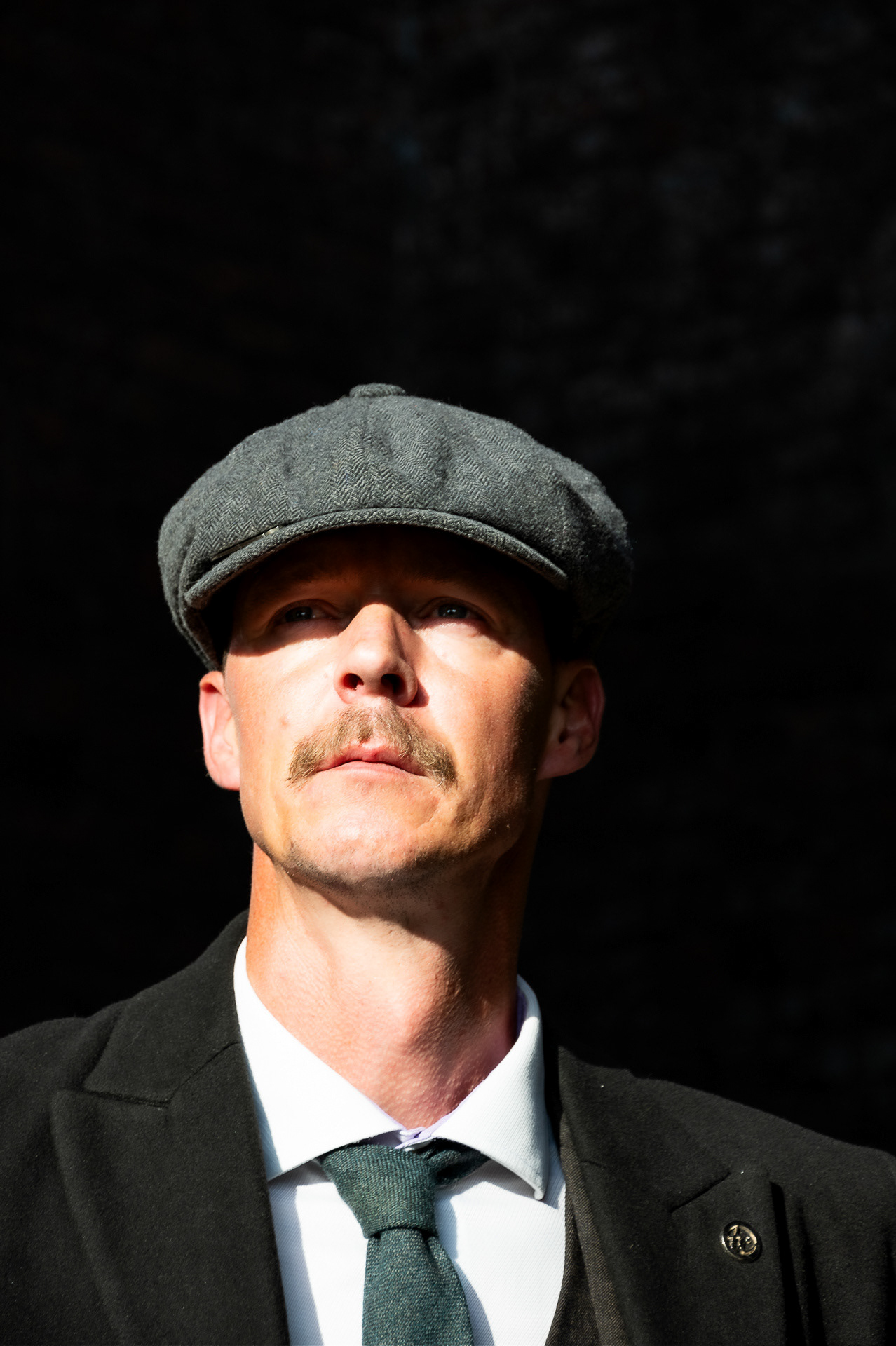 Peaky Shoot - A man dressed in a Jacket shirt and tie , he is also wearing a flat cap. He has a moustache and is looking off to the right  