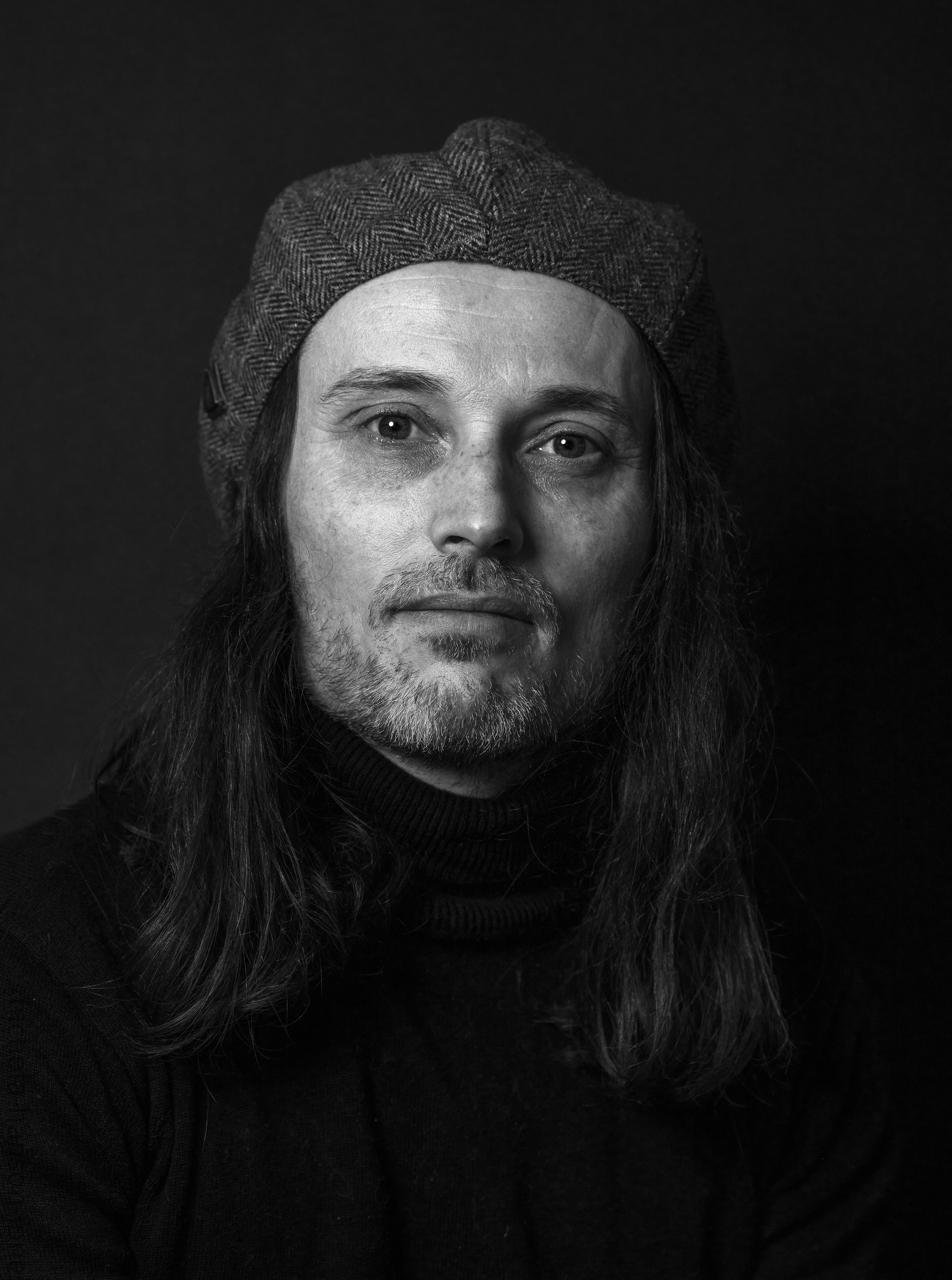 Ian - A man looks straight at the camera, he has a flat cap on that he wears backwards, he has long hair