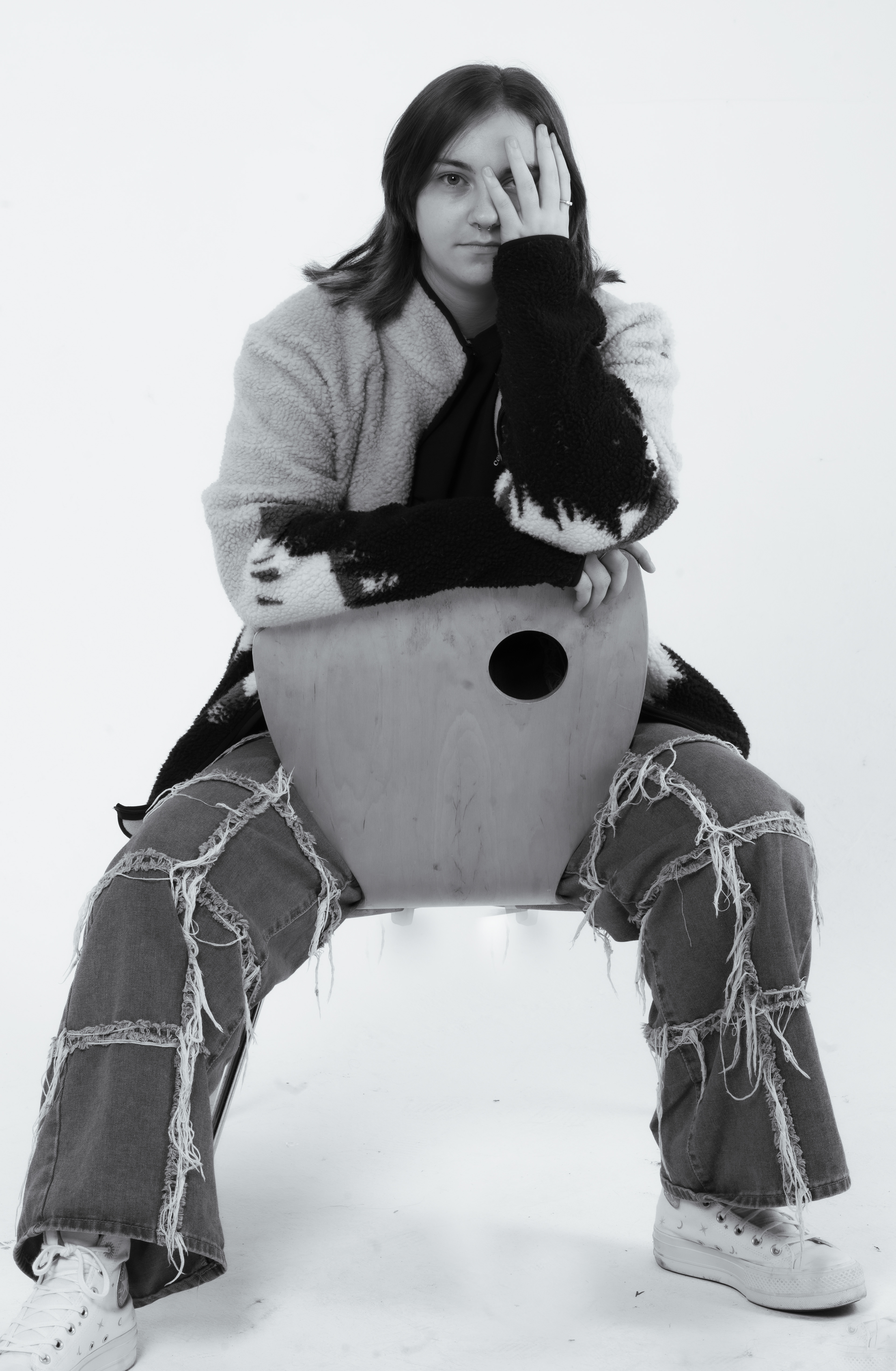 Cassidy -Cassidy - A young woman sits backwards on a chair, she has one arm resting across the back of the chair and the other is held up so that her hand covers her left eye. She wear jeans a sweat shirt and trainers. She is looking straight at the camera with the uncovered eye