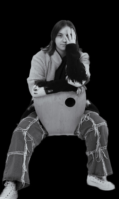 Women in Photography Exhibition 2024 - A young woman sits backwards on a chair, she has one arm resting across the back of the chair and the other is held up so that her hand covers her left eye. She wear jeans a sweat shirt and trainers. She is looking straight at the camera with the uncovered eye 