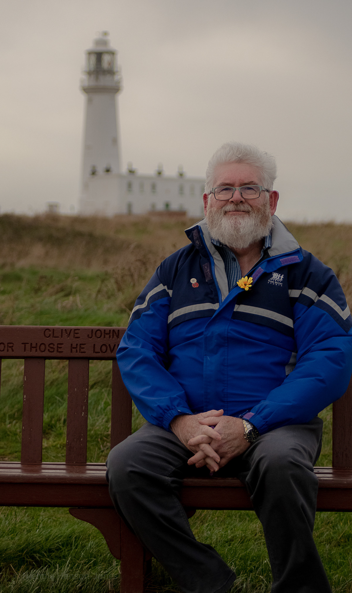 A man is sat on a bench behind him there is a grassy bank with a lighthouse on the top