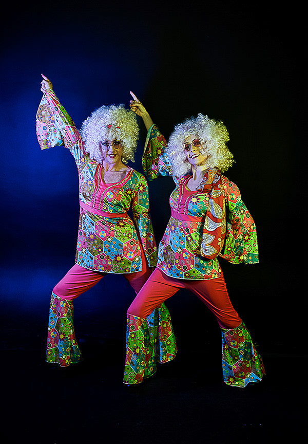 Code Red Promo Shoot - two women dressed in 1970's style clothing which is brightly coloured they are also wearing blond curly wigs and sunglasses, they are pointing upwards with their right arms while their left is pointing down, there stance is wide and they are looking at the camera and smiling  
