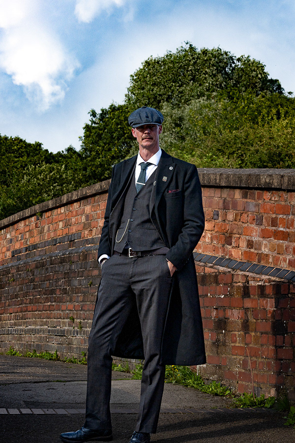 Peaky Shoot A man wearing a long coat a waistcoat and shirt and tie he has his hands in his pockets and is stood outside, there is brickwork and trees behind him, he looks straight at the camera