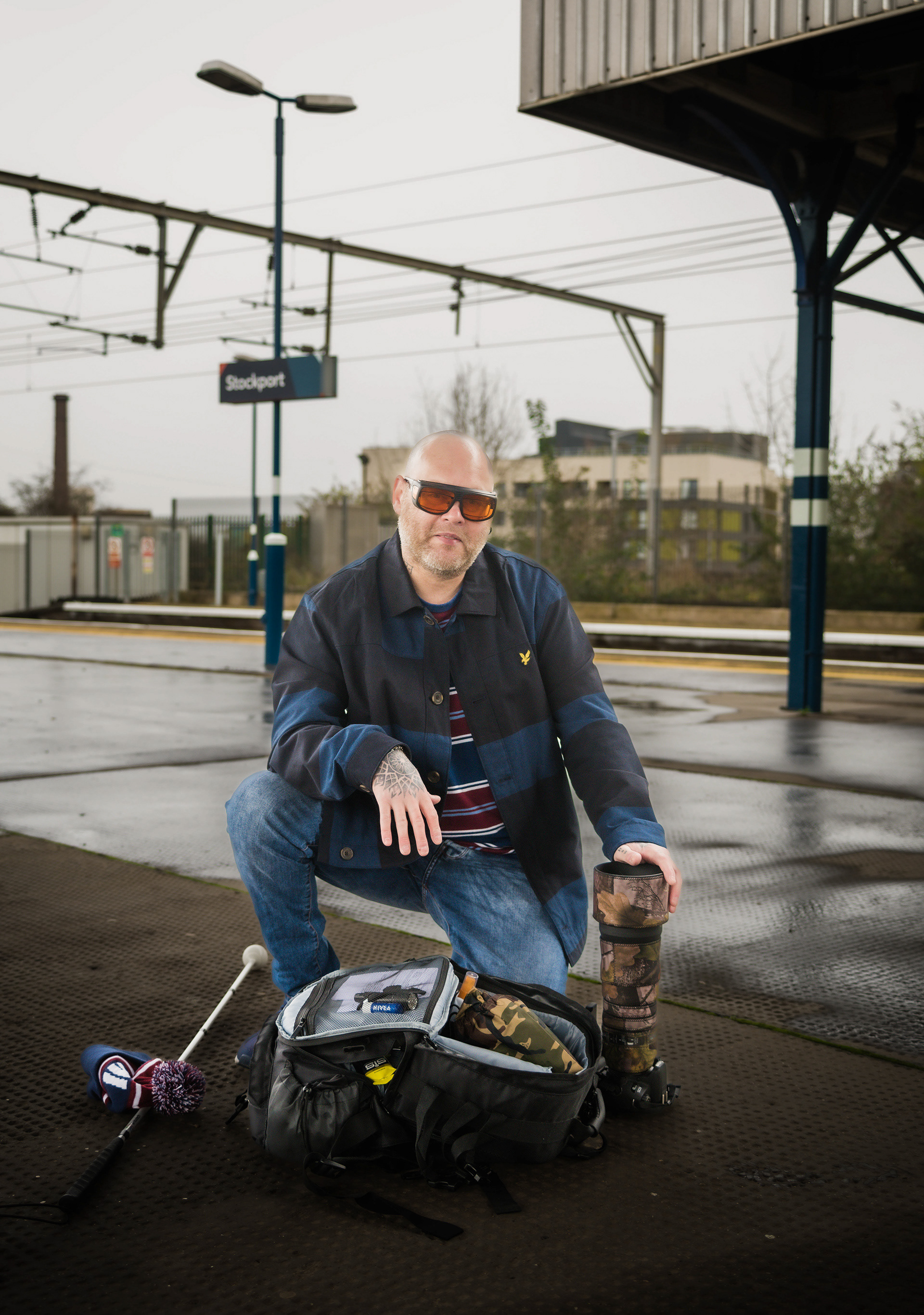 A man kneels down on the platform of a train station. by his side is a white Caine, he wears a pair of yellow coloured glasses there is a bag in front of him and he has his one hand placed on a long lens
