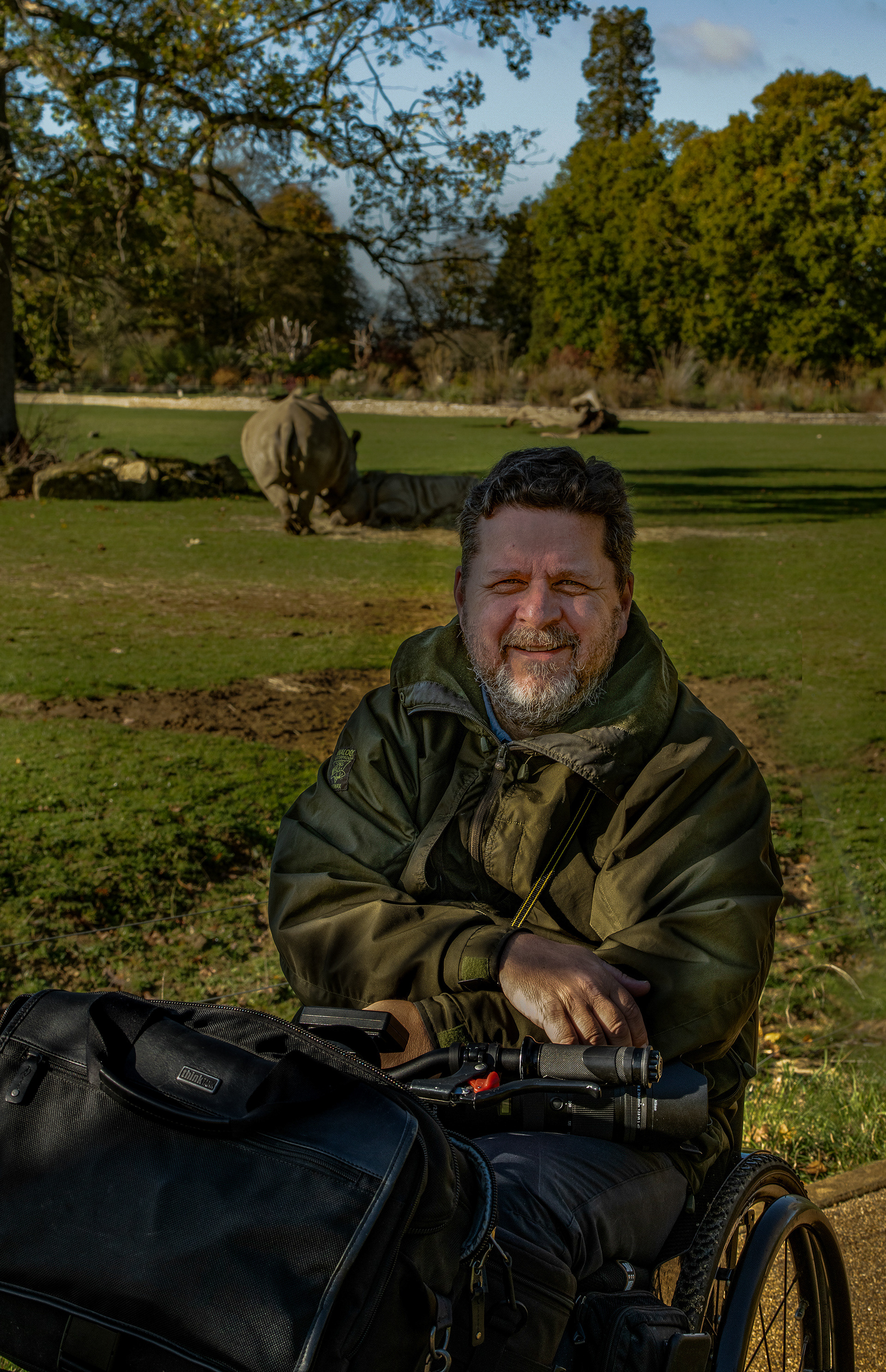 A man sits in a wheelchair, he is in front of an enclosure with a rhino in the background