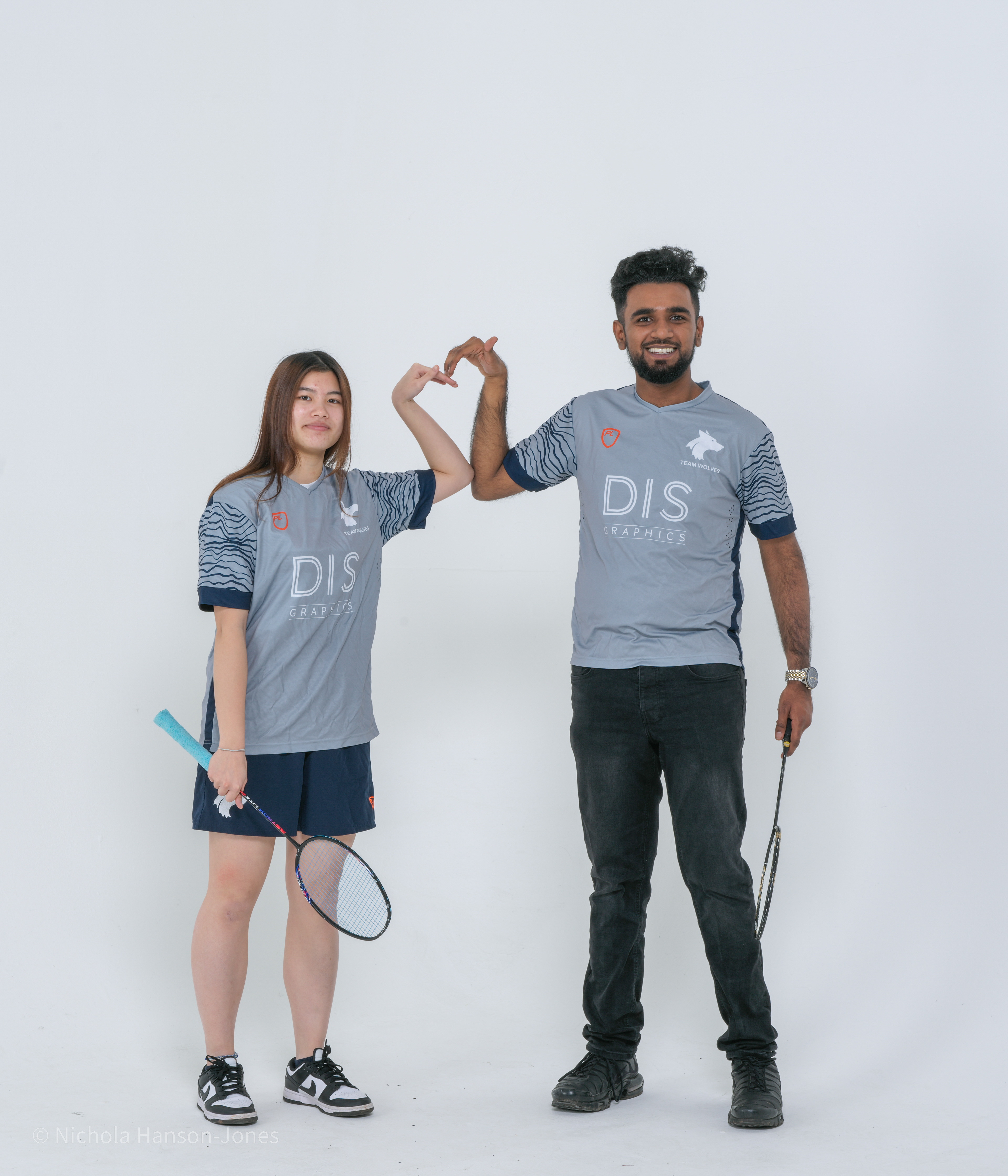 A young man and women wearing sports wear and holding badminton rackets, they also have put their arms together to make a heart shape.