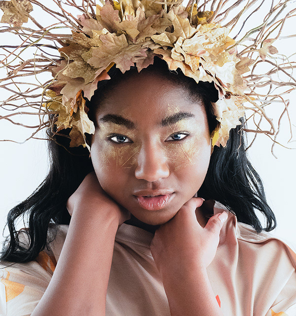 Niah Model shoot - A black woman looking at the camera, her hands are placed gently under her chin, she has gold patterns on her face and wears a gold head dress made form leaves and twigs 